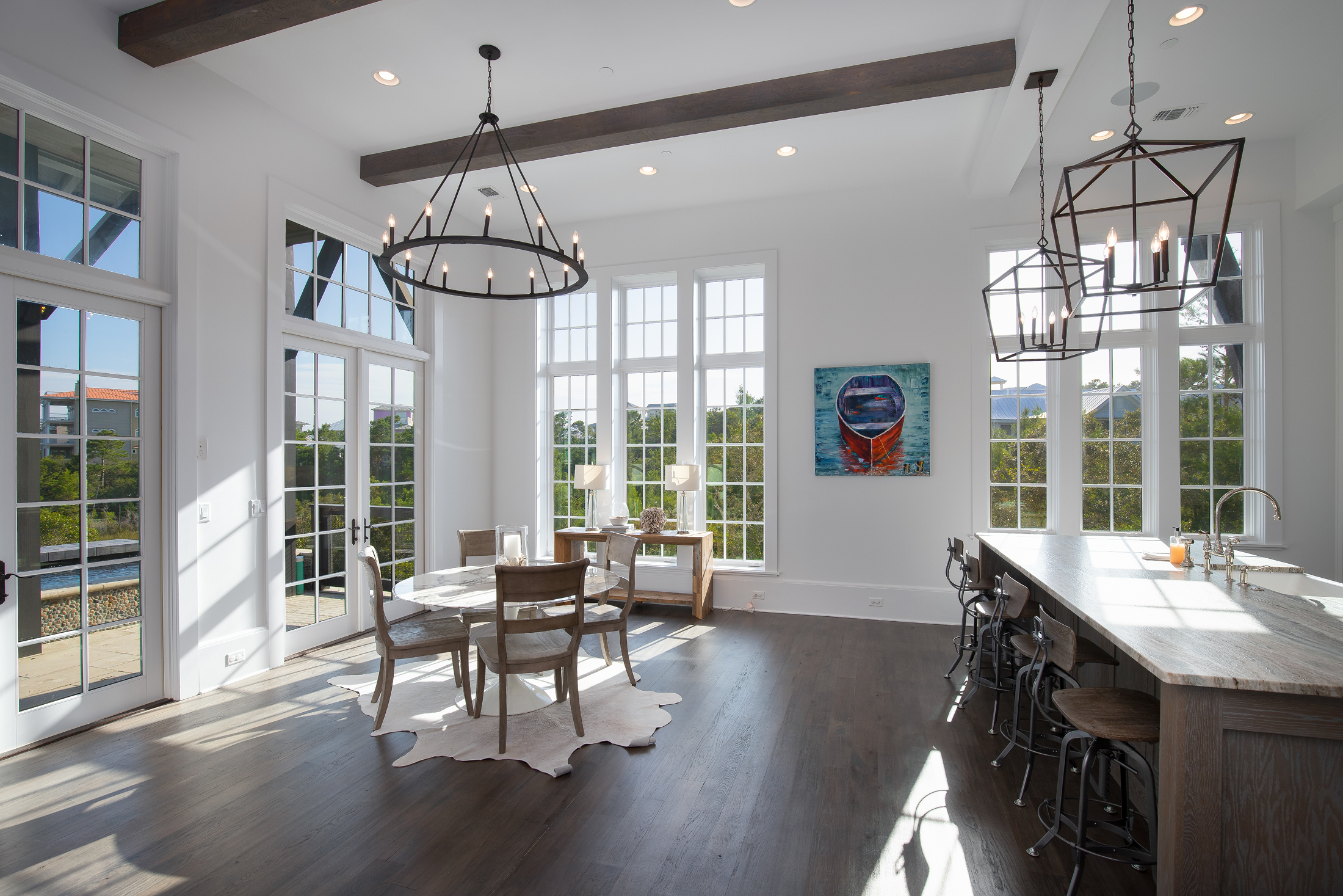 Bright dining area with large windows, a wooden dining table with four chairs, and a kitchen bar with four stools, pendant light fixtures, and a colorful artwork on the wall.