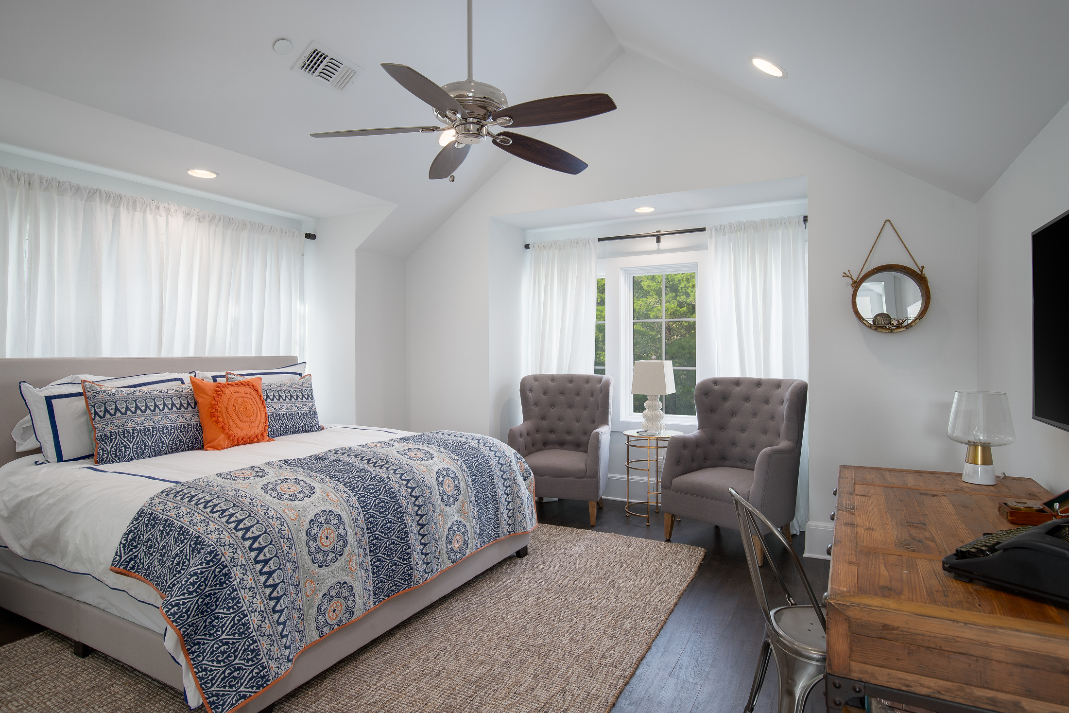 Bright bedroom with a bed that has a blue and white patterned quilt and orange pillows. Two gray tufted armchairs by a window with white curtains. Small side table with a lamp. Wooden desk with a lamp and a TV mounted on the wall.