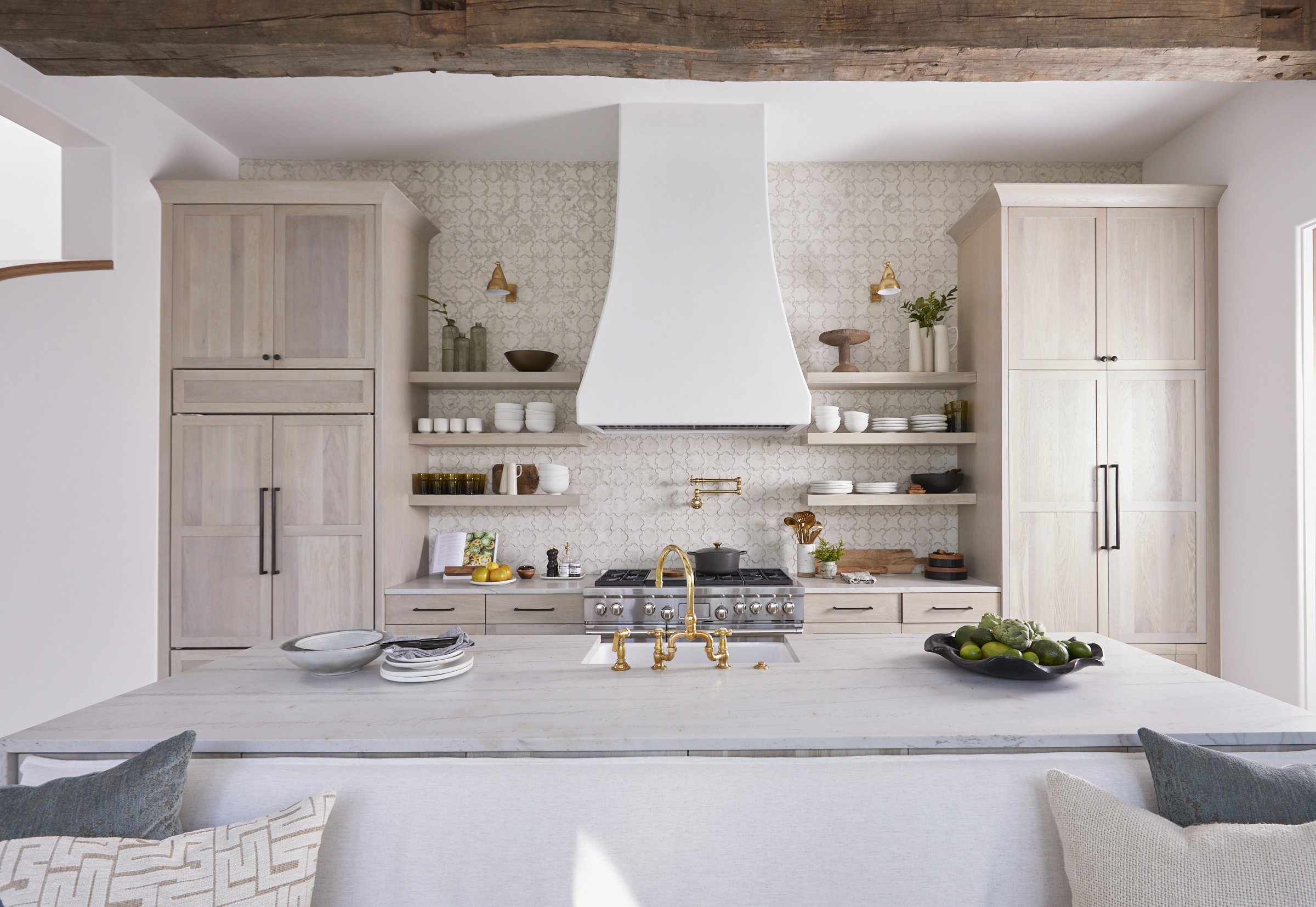 A bright, modern kitchen with a white island, gold faucet, open shelves with dishes, and light wood cabinetry.