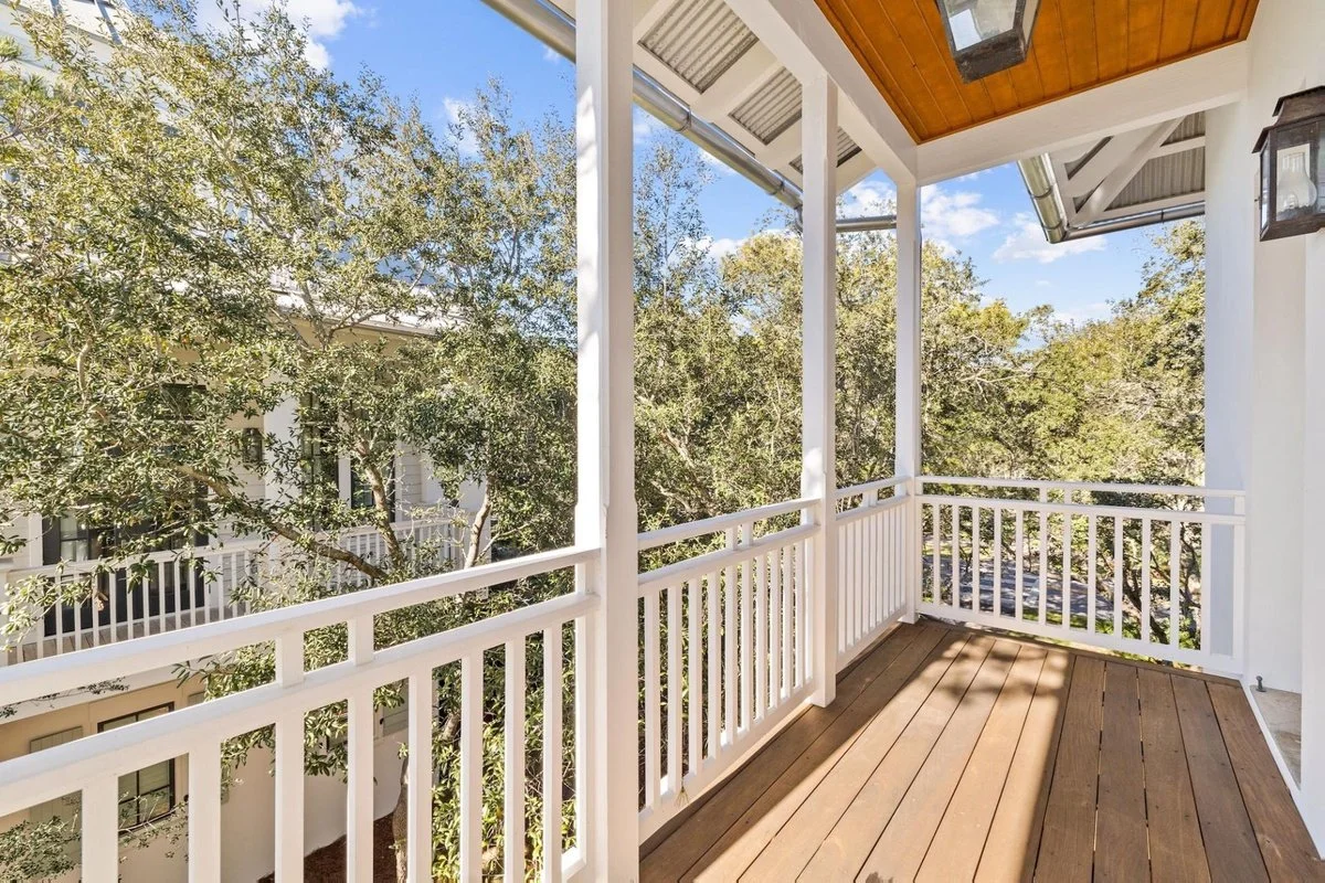A wooden balcony with white railings, overlooking leafy trees and neighboring buildings, under a partly cloudy sky.