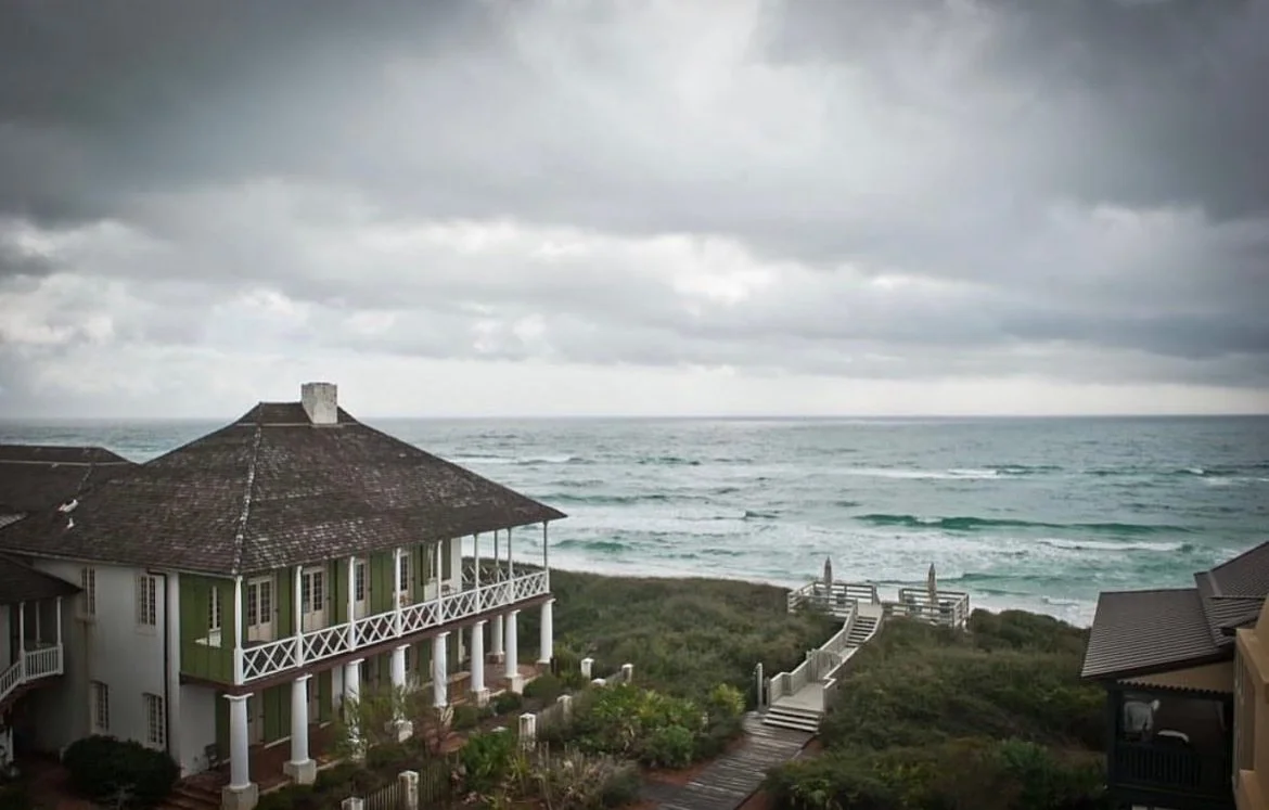 A house with a green exterior and a balcony facing the ocean, with stairs leading down to a beach area, under a cloudy sky.