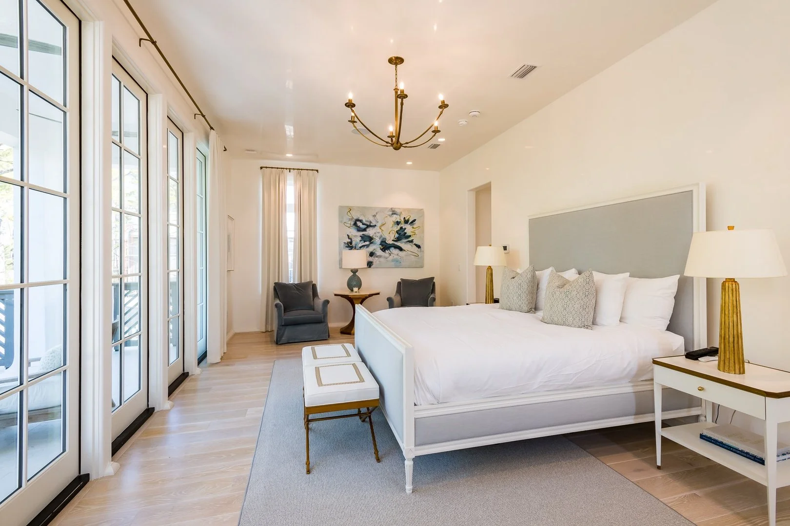 A spacious, bright bedroom with a large bed, white bedding, and multiple pillows. On either side are white nightstands with gold lamps. At the foot of the bed is a small bench with gold legs. To the left are multiple glass doors with white curtains, 