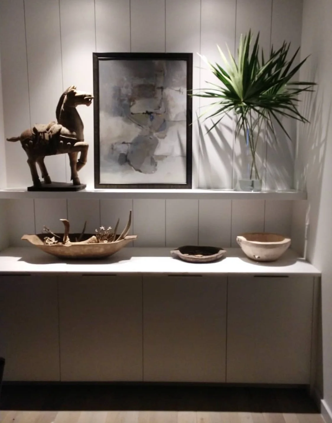 Decorative display with carved wooden horse, ceramic bowls, a framed abstract painting, a large potted plant, and wooden art pieces on a white cabinet.