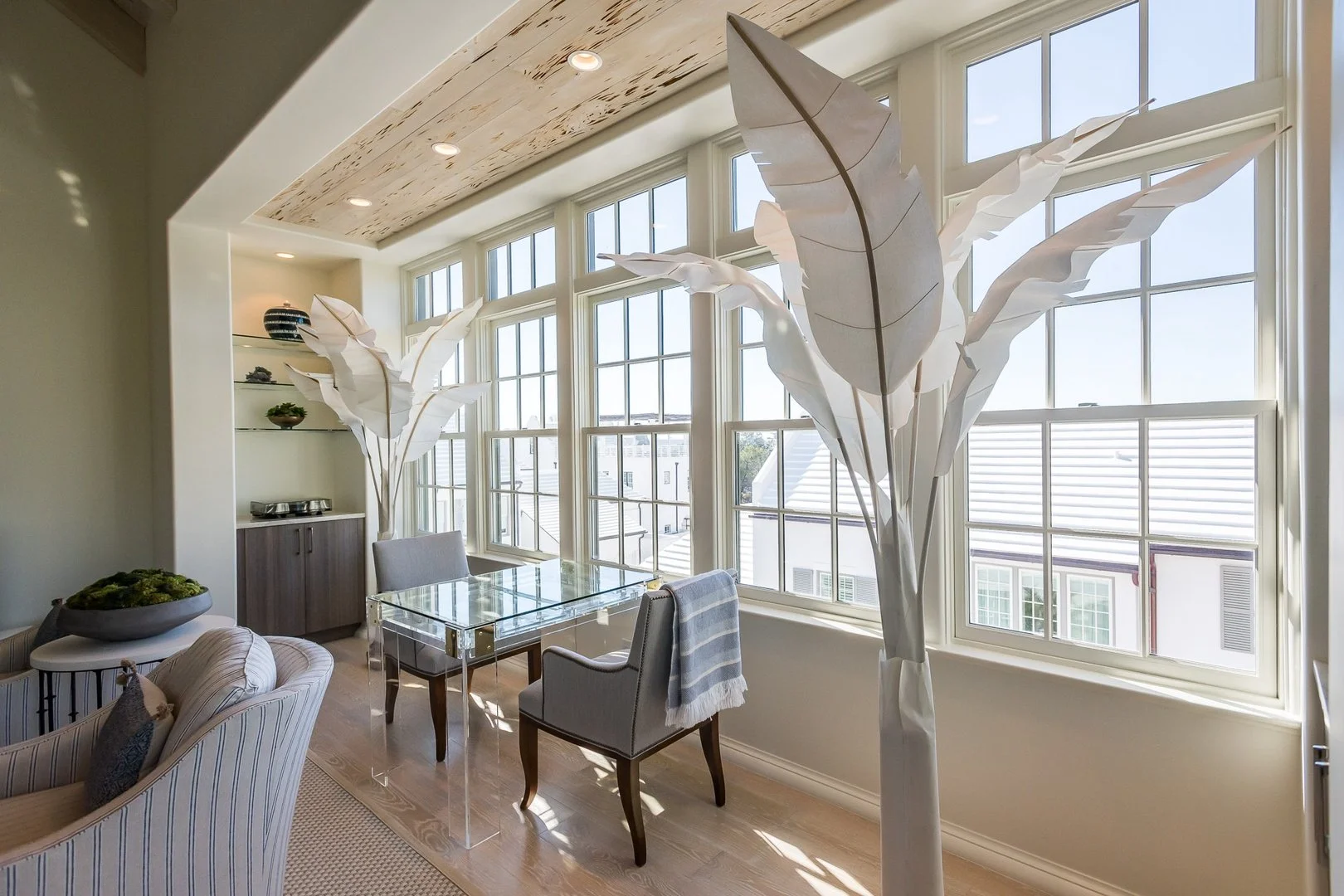Bright living room with large windows, decorative paper trees, a glass dining table, and armchairs.