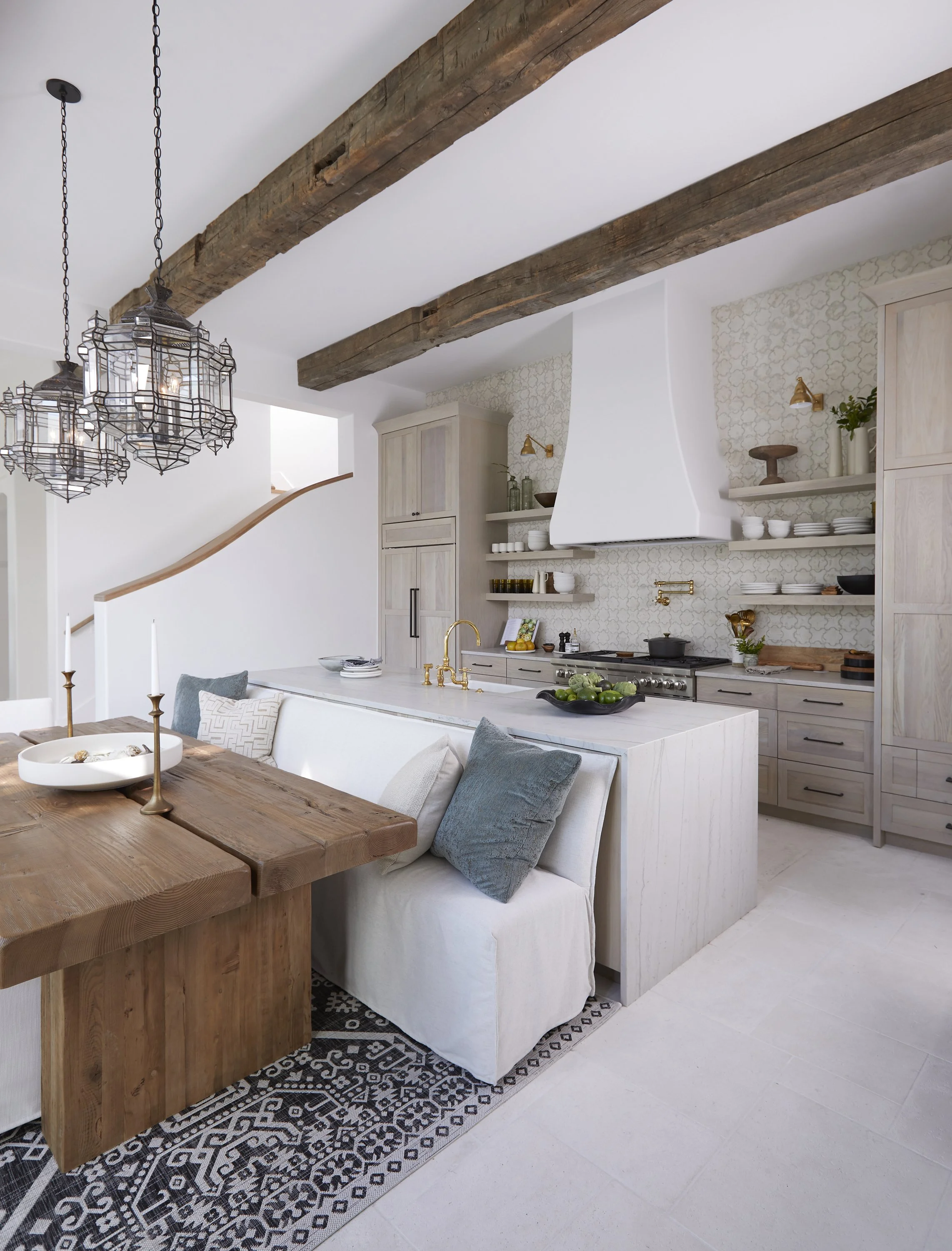 Modern kitchen with white cabinets, open shelves, a white range hood, wooden beams on the ceiling, and a wooden dining table with candles and pillows.