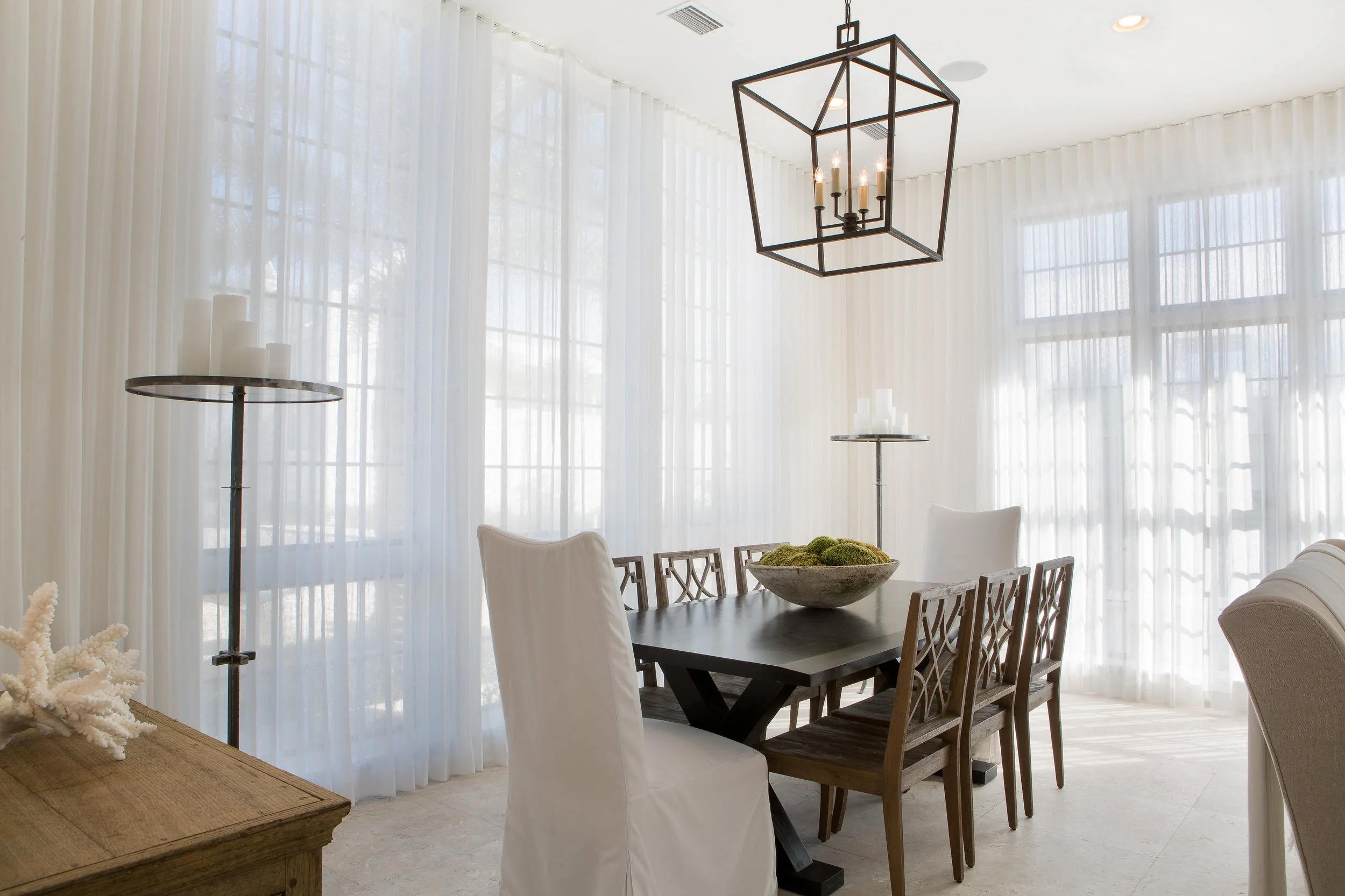 A dining room with sheer white curtains, a black dining table with six chairs, a decorative bowl with moss, tall candle holders with candles, and a modern geometric chandelier hanging from the ceiling.