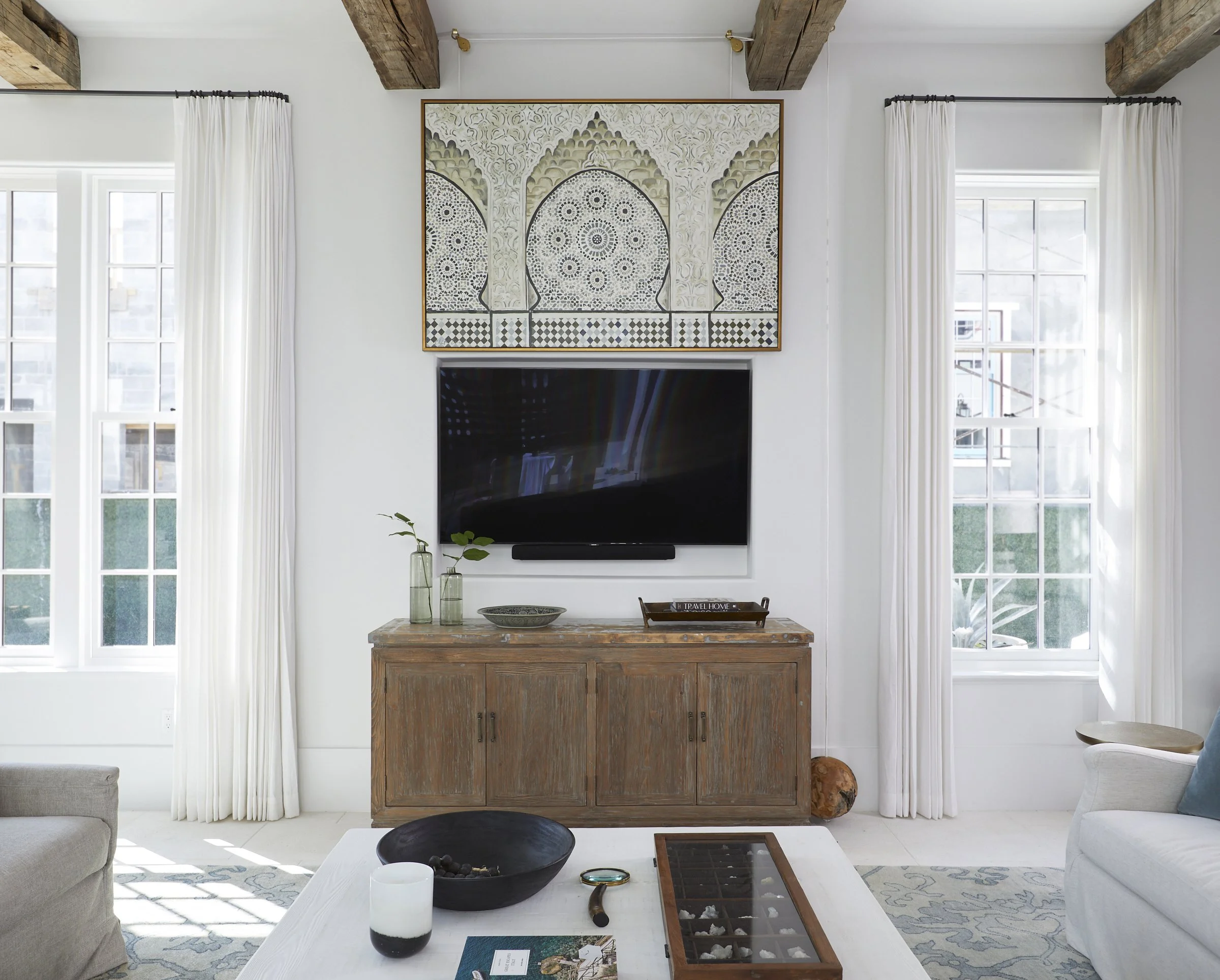 Living room with white walls, large windows with white curtains, a flat-screen TV mounted on the wall, and a wooden cabinet below it. Decorative items and greenery are on the cabinet, and artwork hangs above the TV. A coffee table with various items 