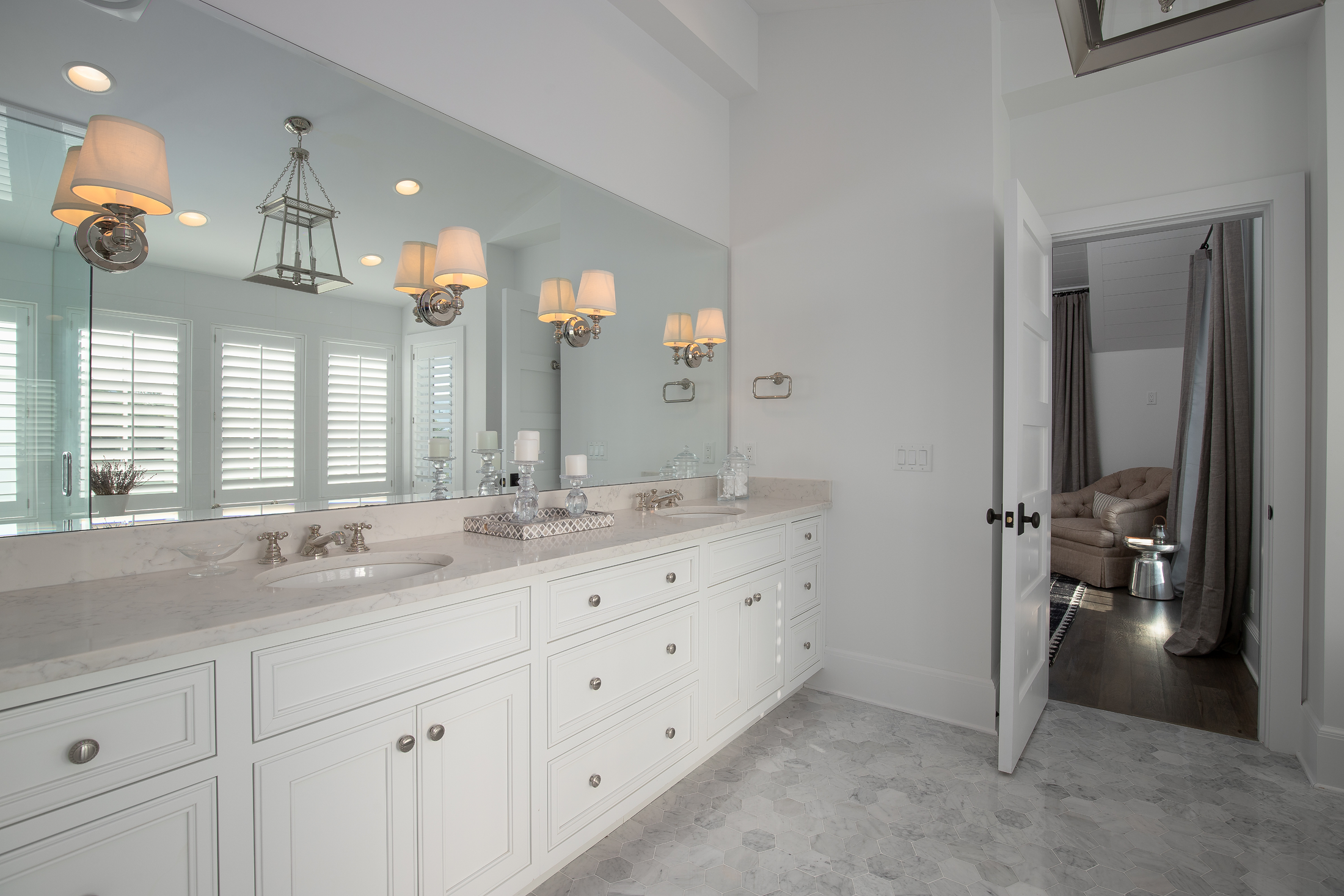 A bright bathroom with white cabinetry, a large mirror, marble countertop, and four elegant wall sconces. The floor is covered with gray and white hexagonal tiles. There are blinds on the windows reflected in the mirror, and an open door leads to a r
