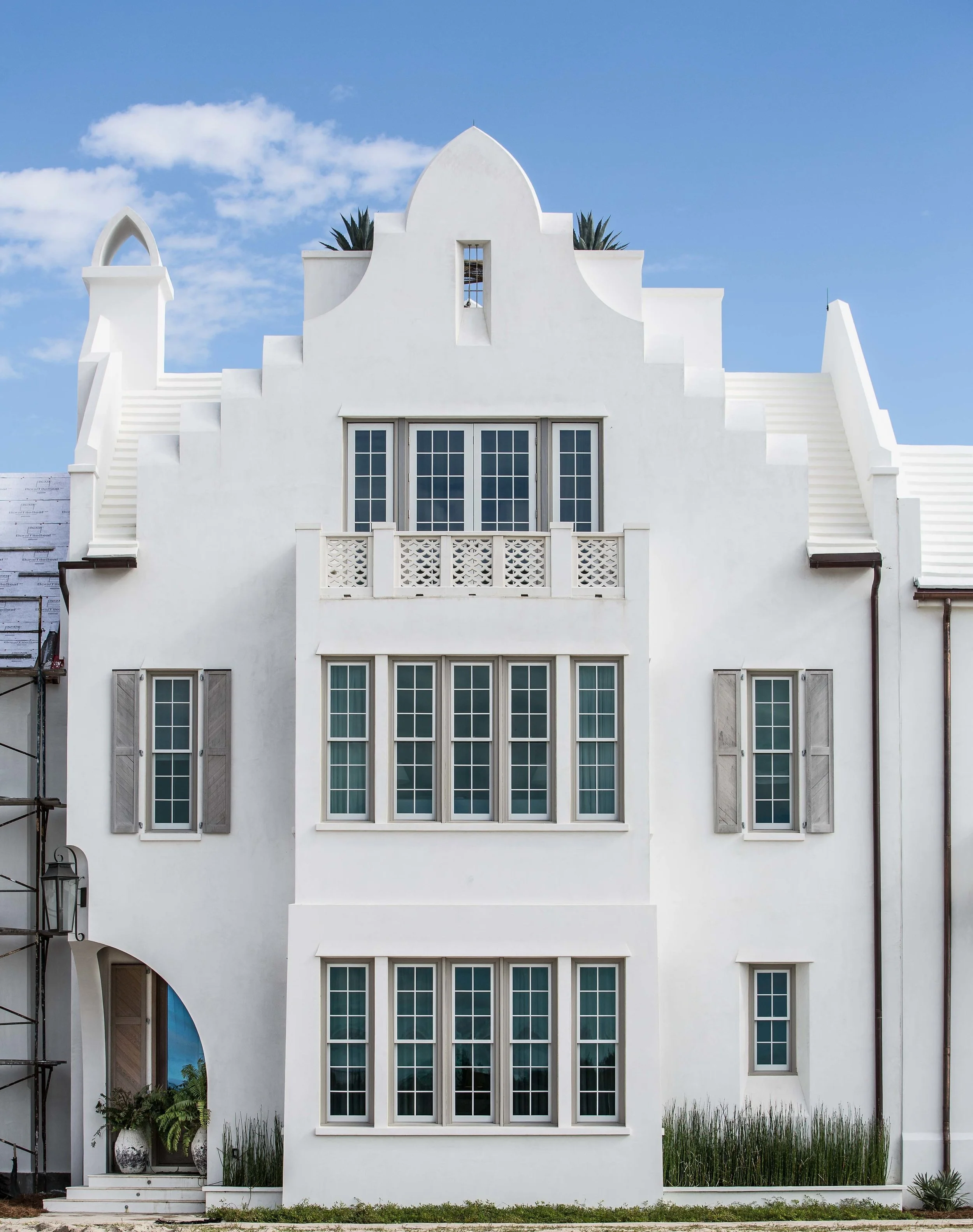 White multi-story house with arched rooflines, multiple windows with gray trim, and a small balcony. Clear blue sky with a few clouds in the background.