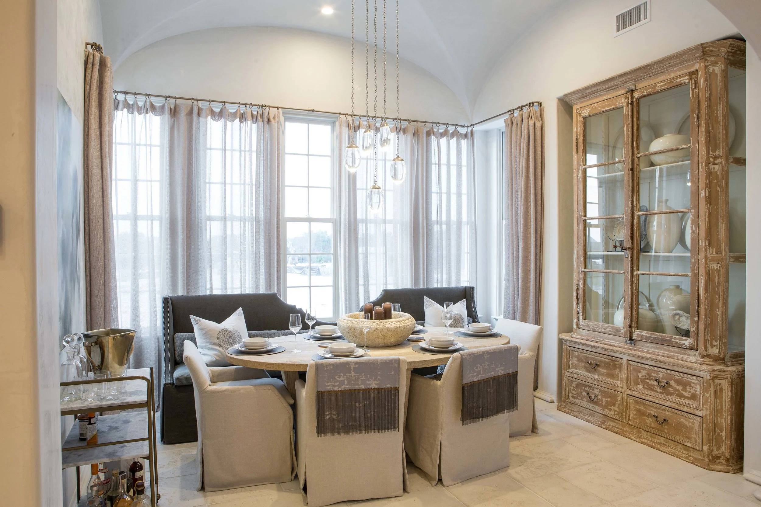 A cozy dining room with a round table set with dishes, glasses, and candles, surrounded by cream and grey upholstered chairs, large windows with sheer curtains, a vintage wooden display cabinet with pottery, and a contemporary chandelier hanging from
