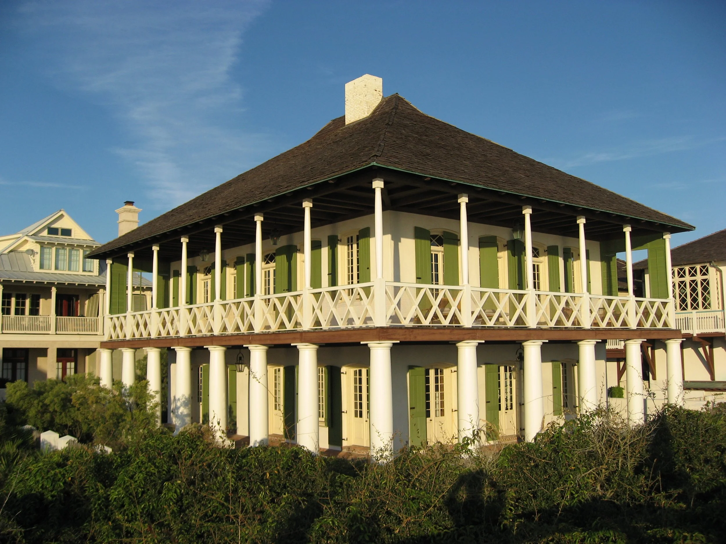 Two-story house with green shutters, white balcony railing, and white columns, under a shingled roof, with neighboring houses and bushes in front, under a blue sky.