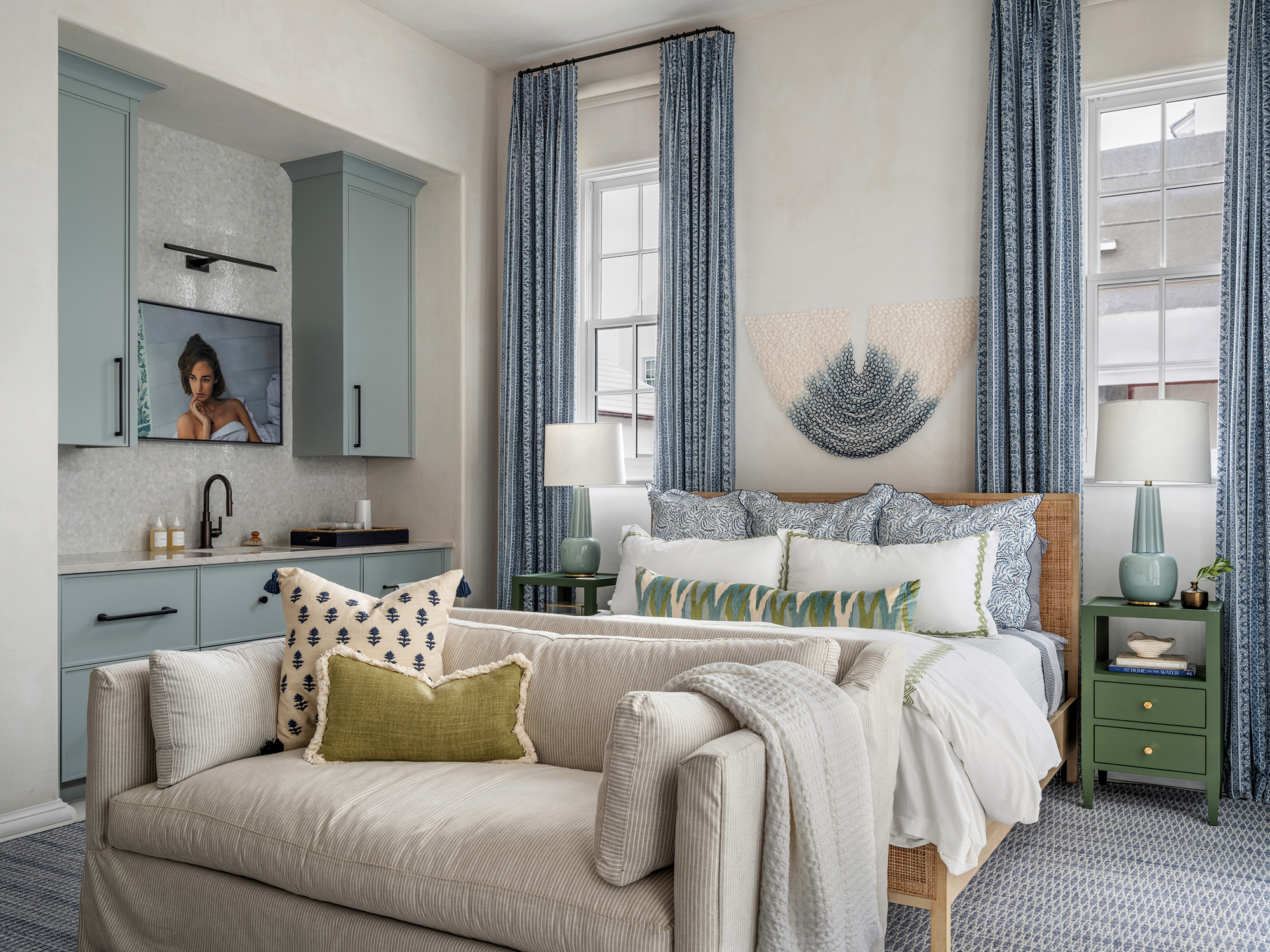 A cozy bedroom with a bed, pillows, and curtains, with a small green nightstand and lamp, blue curtains, a wall hanging, and a beige sofa with pillows in the foreground.