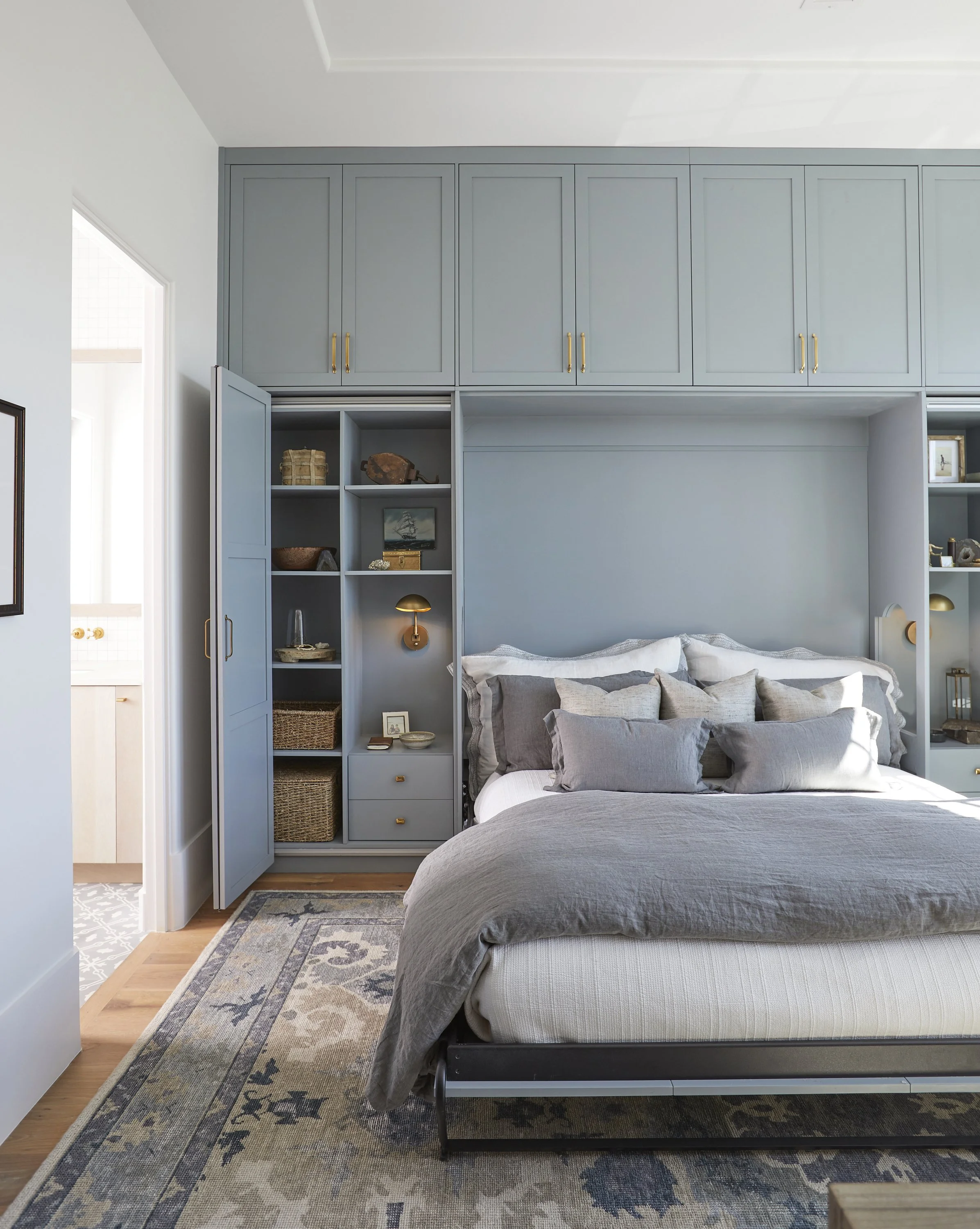 A bedroom with blue built-in cabinets and shelves above a bed, decorated with gray and beige pillows, and a patterned rug on the wooden floor.