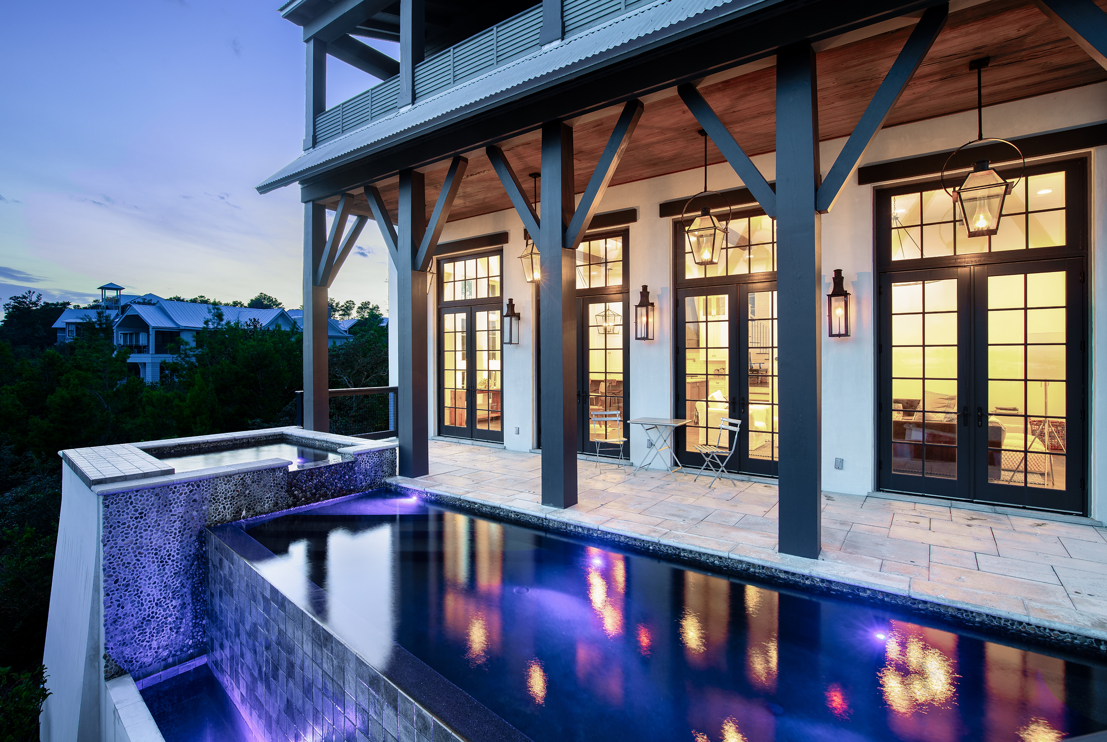 Evening view of a modern house balcony with a small pool and hot tub, illuminated by outdoor lanterns, with a view of neighboring houses and trees in the distance.