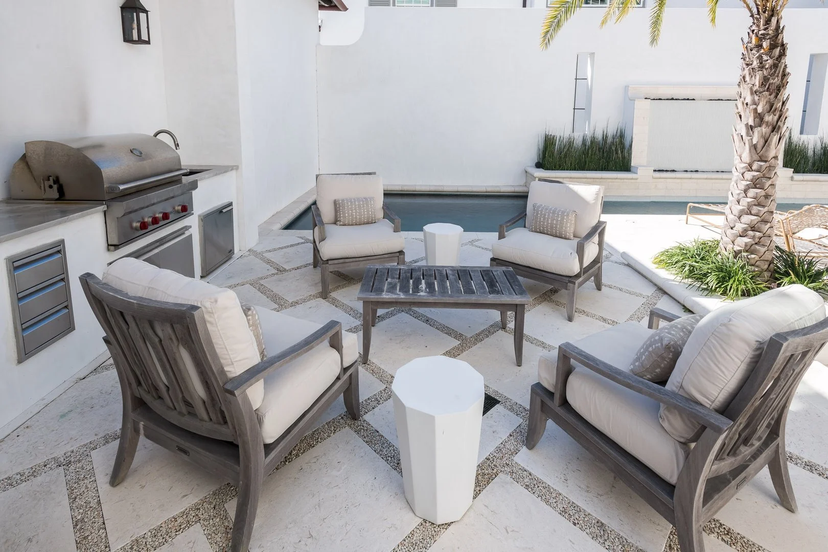 Outdoor patio area with four cushioned wooden chairs around a matching table, a white side table, a built-in grill, a small swimming pool, palm tree, and decorative plants.