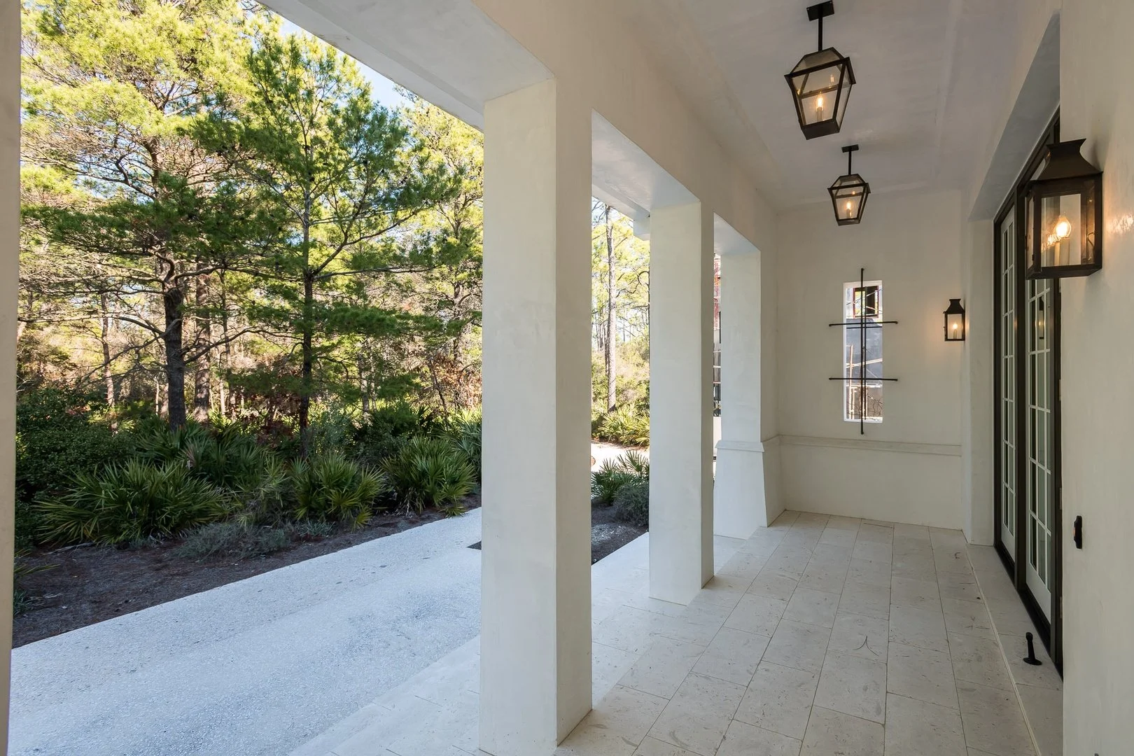 Covered porch with hanging lantern lights, white walls, tiled floor, and a view of trees and shrubbery outside.