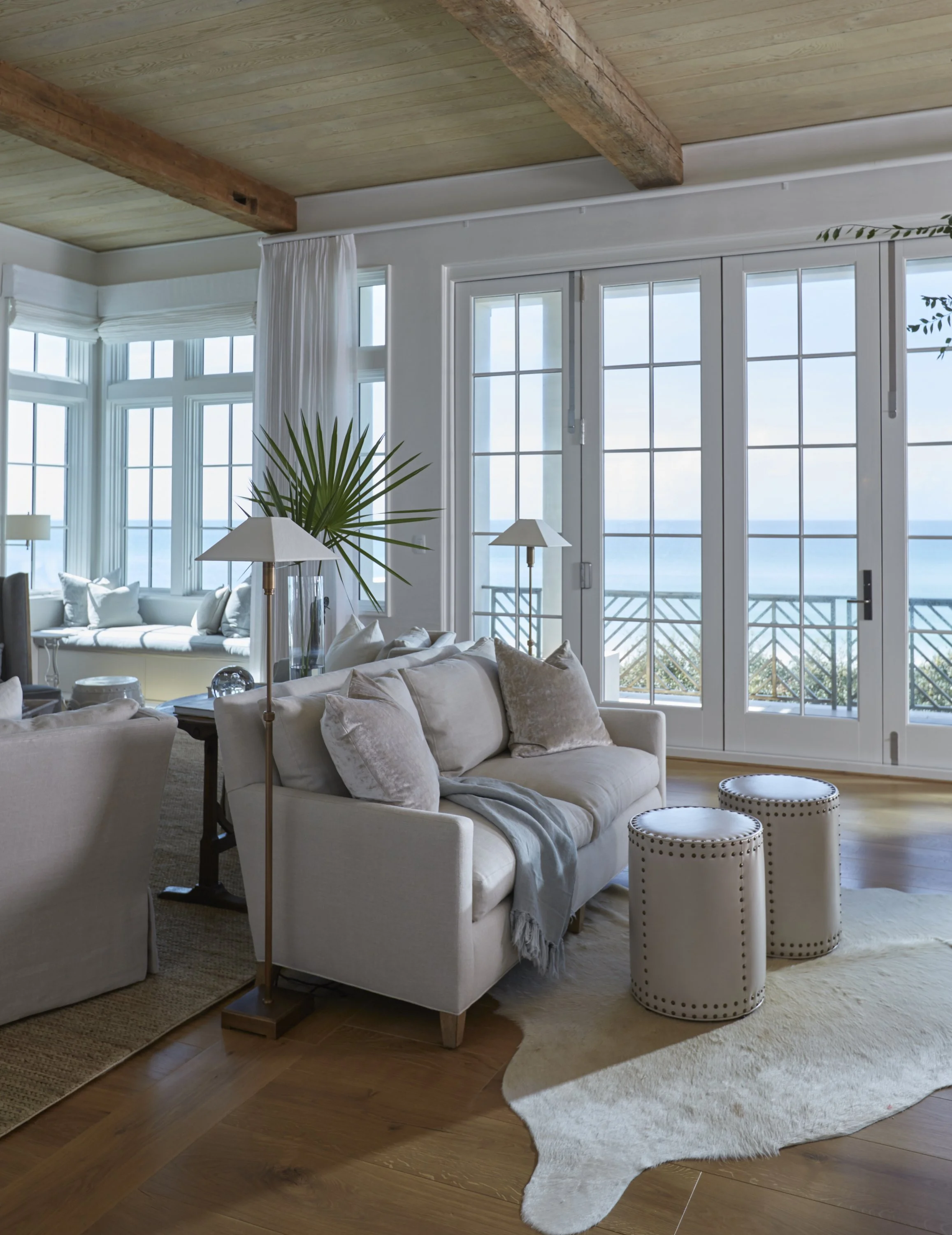 Living room with large windows overlooking the ocean, white sofa with beige pillows, two white stools, green plant, wooden ceiling beams, hardwood floor, and light-colored rug.
