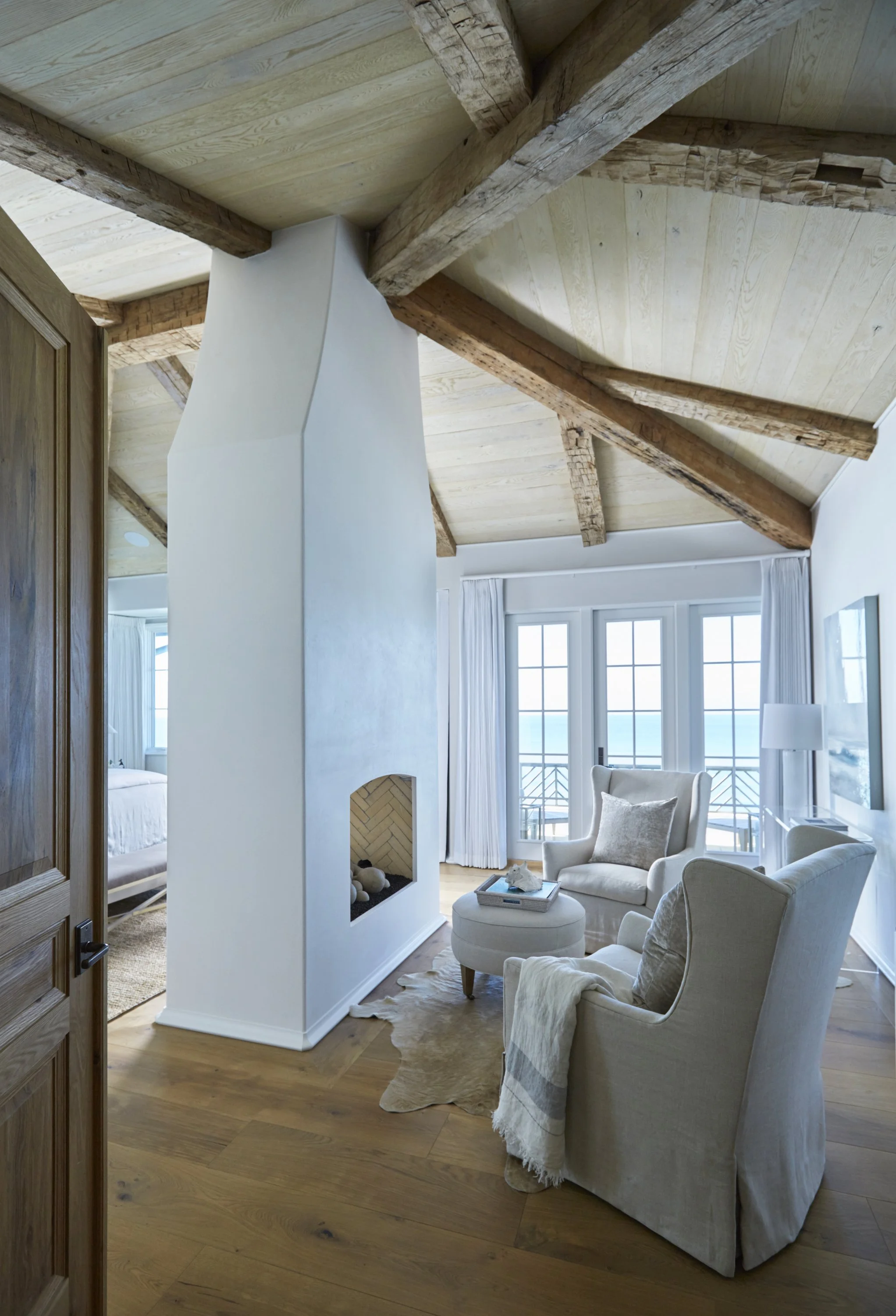 Living room with vaulted wood-beamed ceiling, white fireplace, two armchairs with pillows, ottoman, and large windows with a view of the ocean.