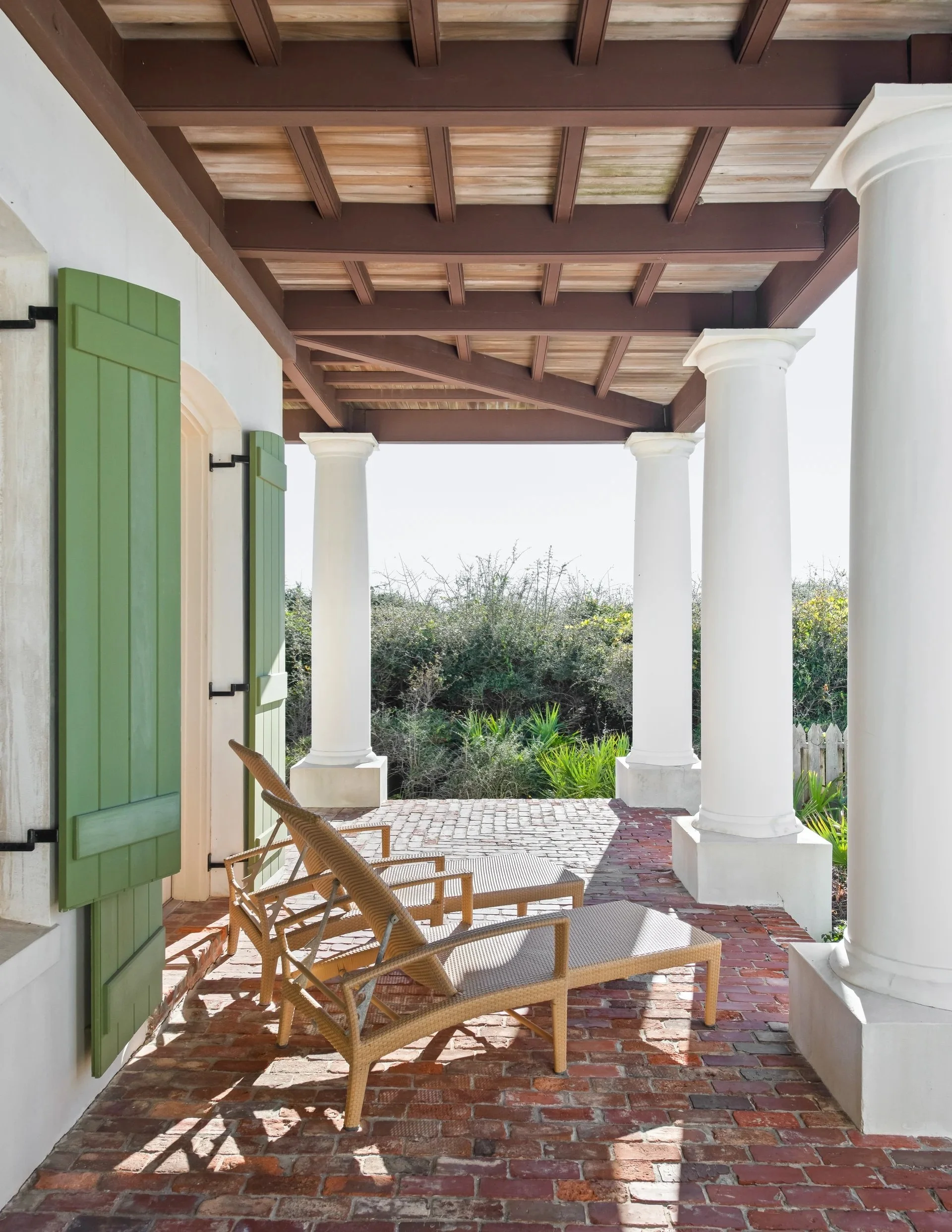 A porch with brick flooring, white columns, and a wooden ceiling, featuring two wicker lounge chairs and green window shutters.