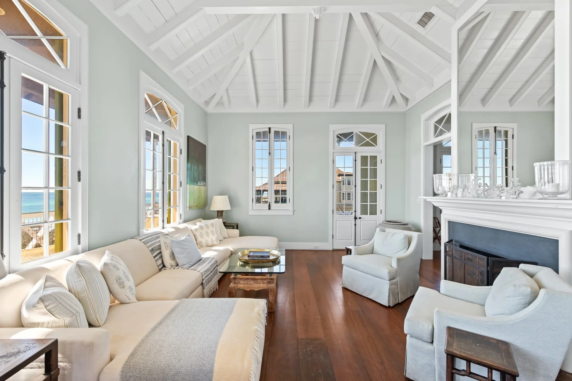 Living room with white walls, high vaulted ceiling, multiple large windows showing ocean view, light-colored furniture, a fireplace, and hardwood floors.