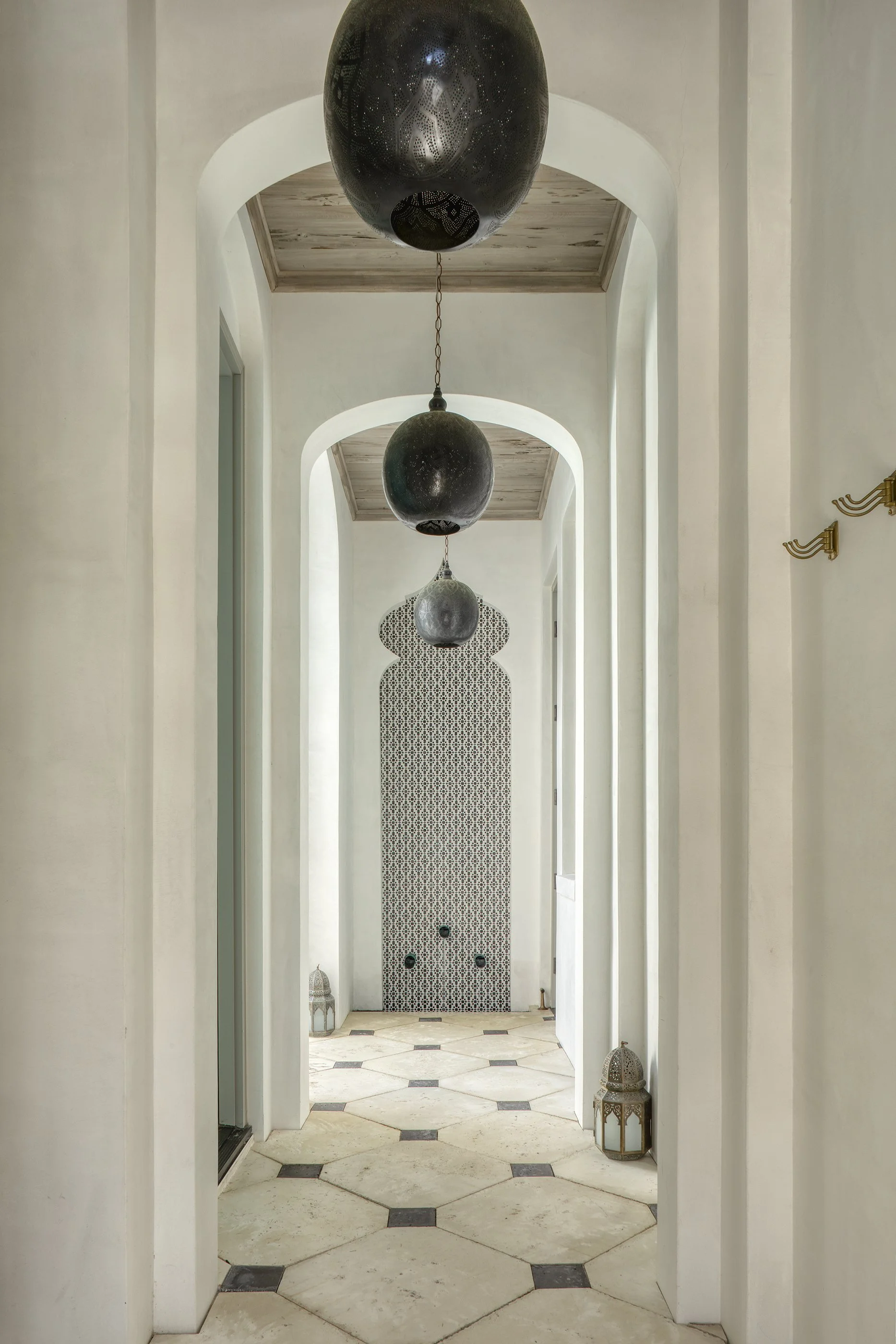 A hallway with white walls, arched doorways, and a tiled floor. Three large, dark pendant lights hang from the ceiling, and decorative lanterns are placed on the floor.