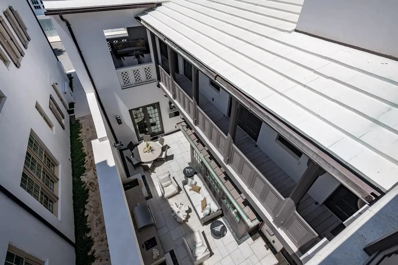 View of a modern outdoor courtyard from above, featuring a round dining table with chairs, lounge seating, and a barbecue grill, surrounded by white walls and multiple windows.