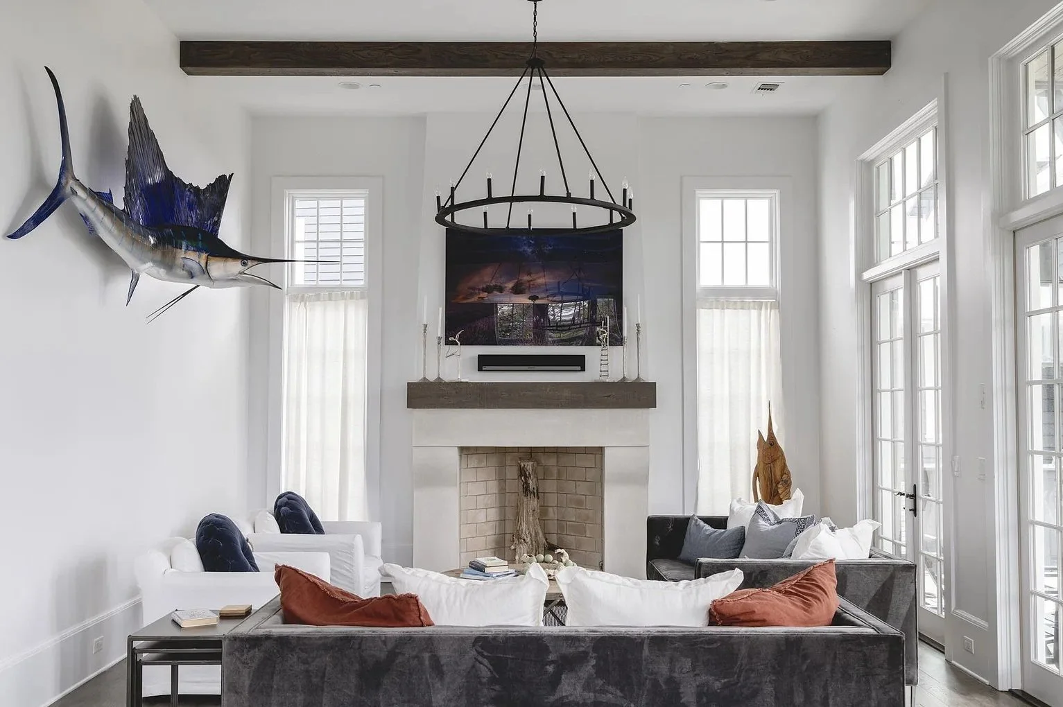 Modern living room with white walls, large windows, and a fireplace. A mounted marlin fish decorates the wall to the left. The room features a black chandelier, various cushions, and a fireplace mantel with decorative objects.