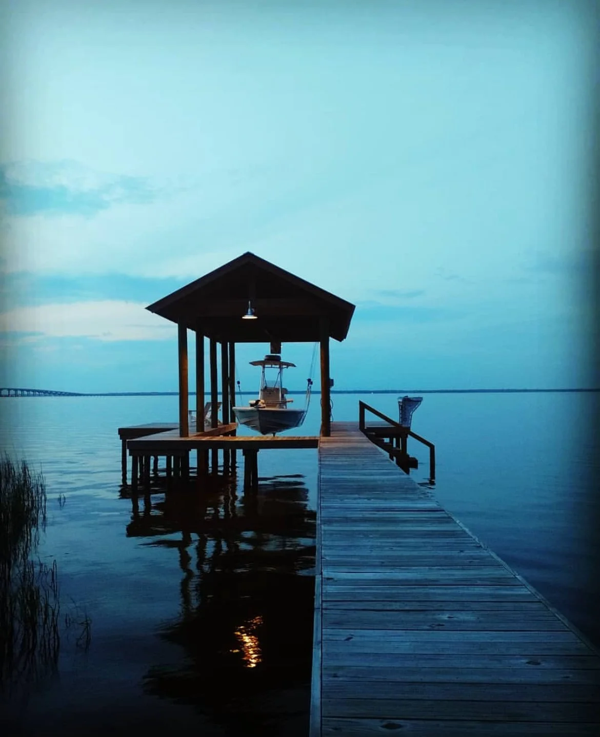A boat dock with a small covered pier extending over calm water, with a boat tied up underneath and a chair on the dock, under a twilight sky.