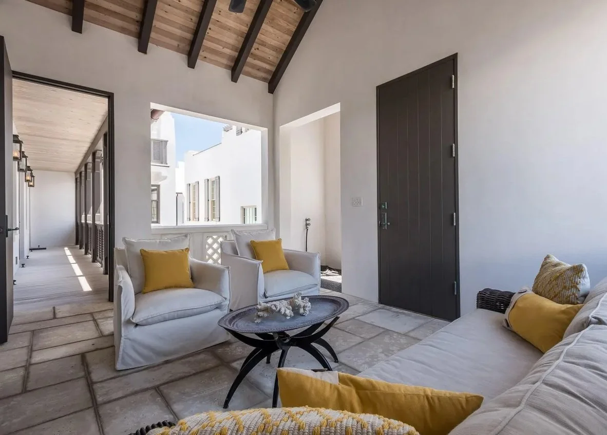 Living room with white sofas and yellow pillows, stone tiled floor, open wall with view of neighboring white buildings, and a dark door.