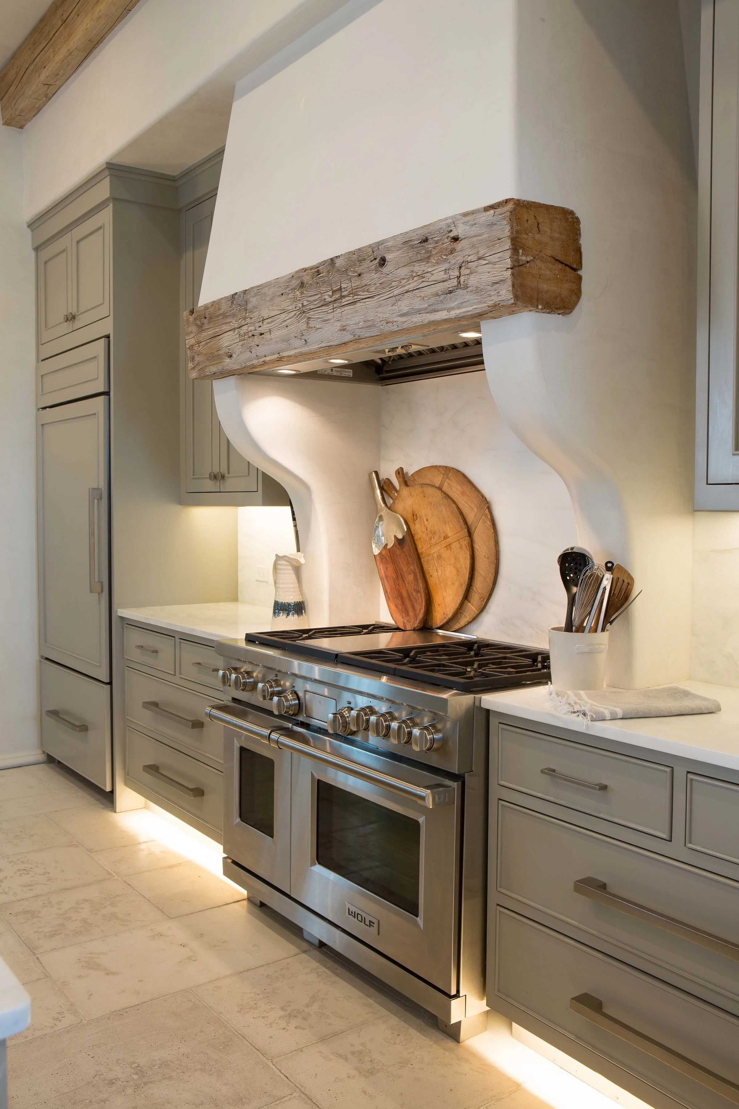 Modern kitchen with a stainless steel Wolf stove, cabinetry, and a rustic wooden beam above the stove, with cutting boards and utensils on the counter.