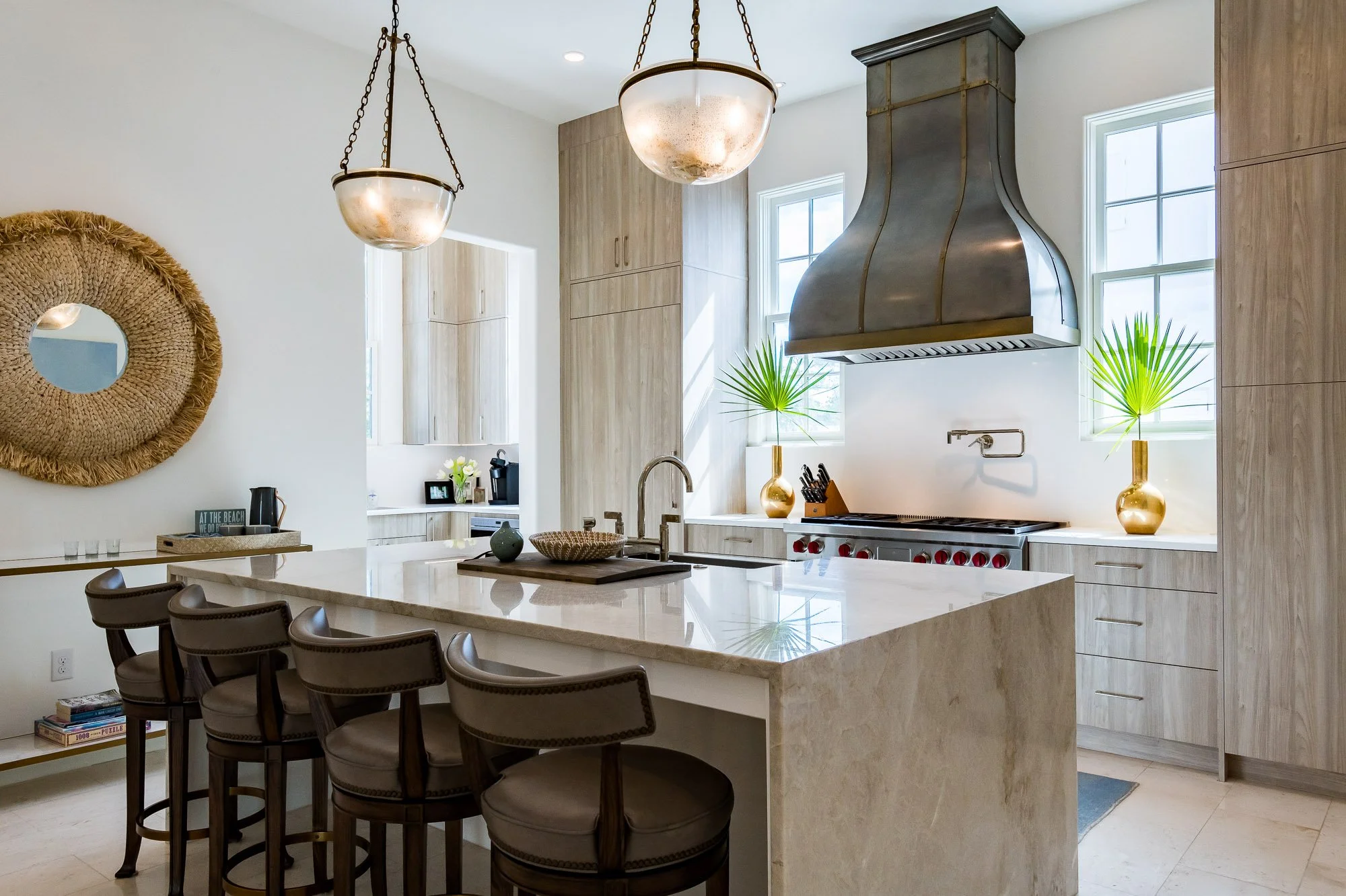 Modern kitchen with an island, four bar stools, a stainless steel range hood, two windows with pendant lights, decorative vases with palm leaves, and wooden cabinetry.