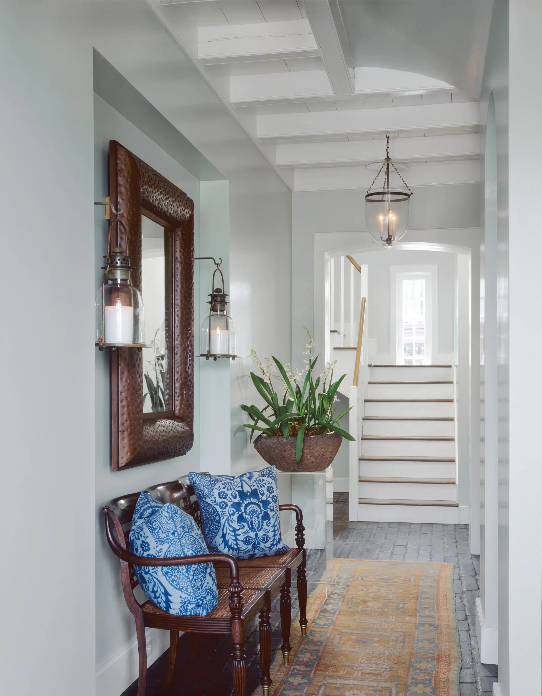 Entryway with a small wooden bench with blue patterned pillows, a large plant in a decorative pot, a mirror with a brown textured frame, wall-mounted lantern-style lights, a hanging ceiling light, white staircase at the end, and a multicolored runner