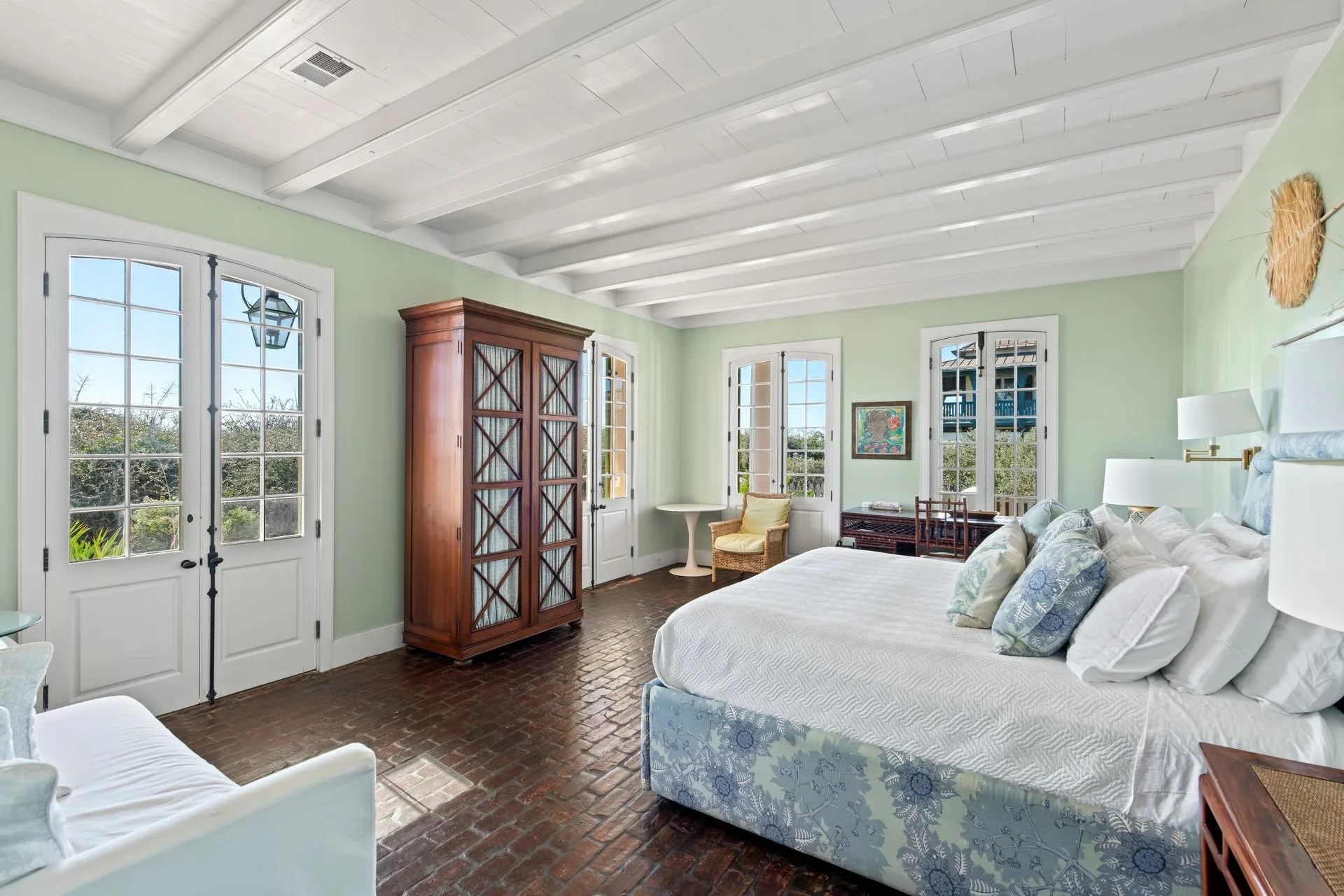 Bright bedroom with large windows, white ceiling with beams, wooden armoire, white bed with multiple pillows, small yellow armchair, and a desk with a chair, overlooking greenery outside.