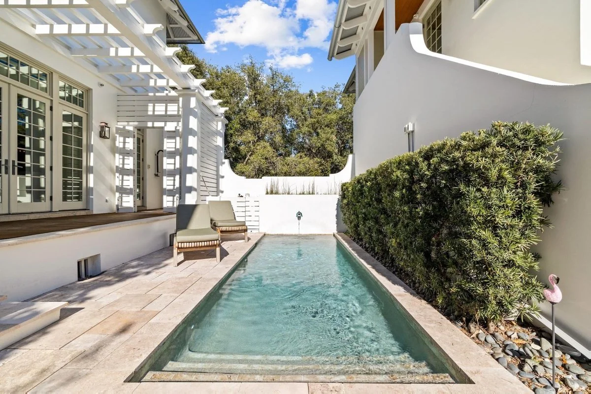 Backyard with a narrow rectangular swimming pool, two poolside chairs, a white wall with a water spigot, green bushes, and a white house with glass doors and a pergola.