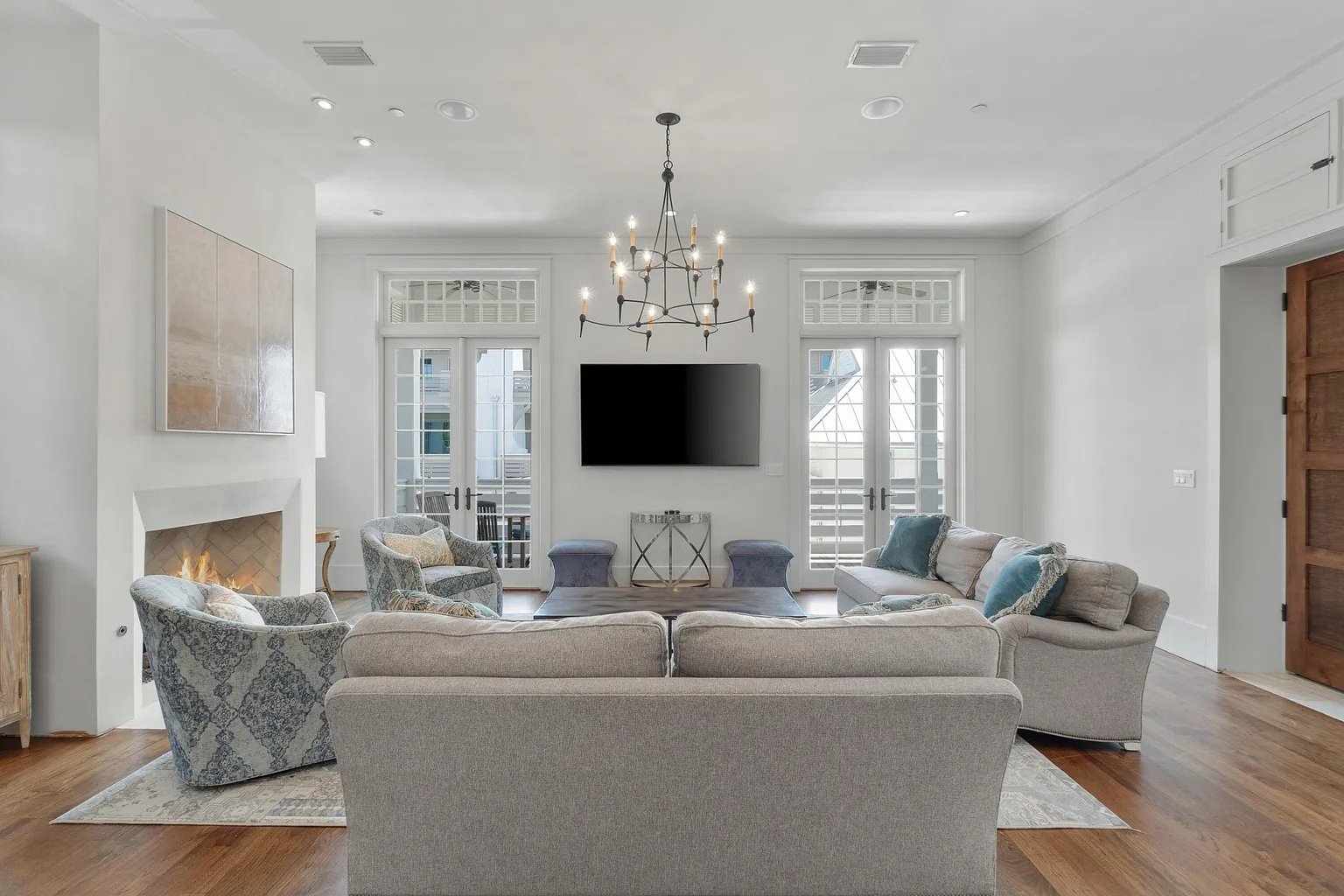 Bright living room with beige sofas and patterned armchairs around a coffee table, fireplace on the left, large flat-screen TV on the wall, and French doors leading to the balcony, decorated in neutral tones with a chandelier hanging from the ceiling