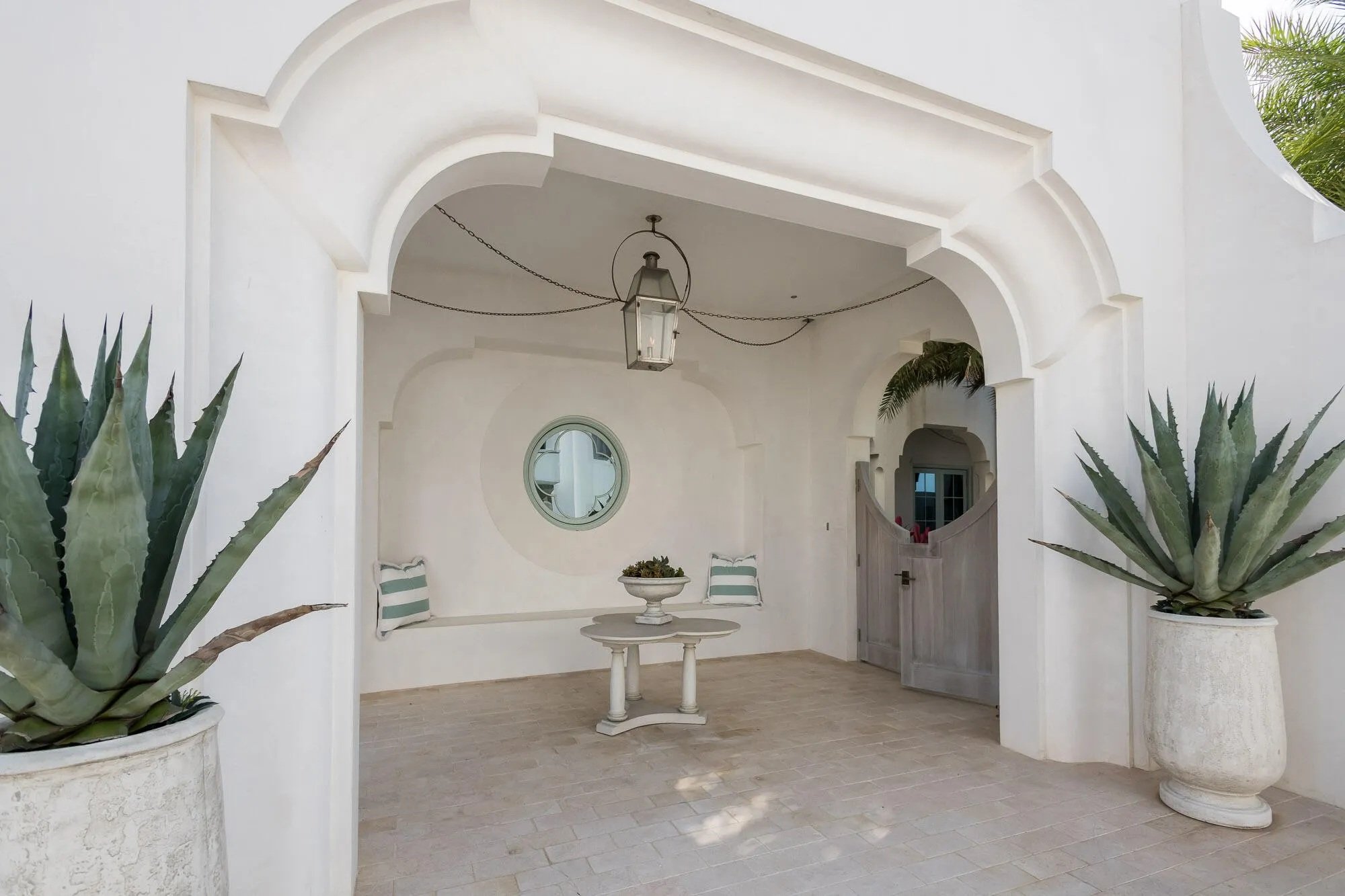 White covered patio with large potted agave plants, a round table with a planter, a bench with cushions, a circular window, and a hanging lantern