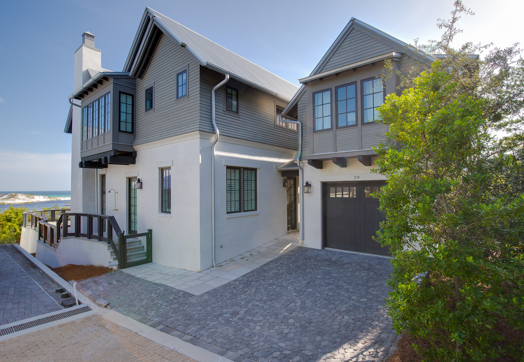 A modern multi-story house near the beach with a black garage door, gray siding, large windows, and a tree in front, under a clear blue sky.