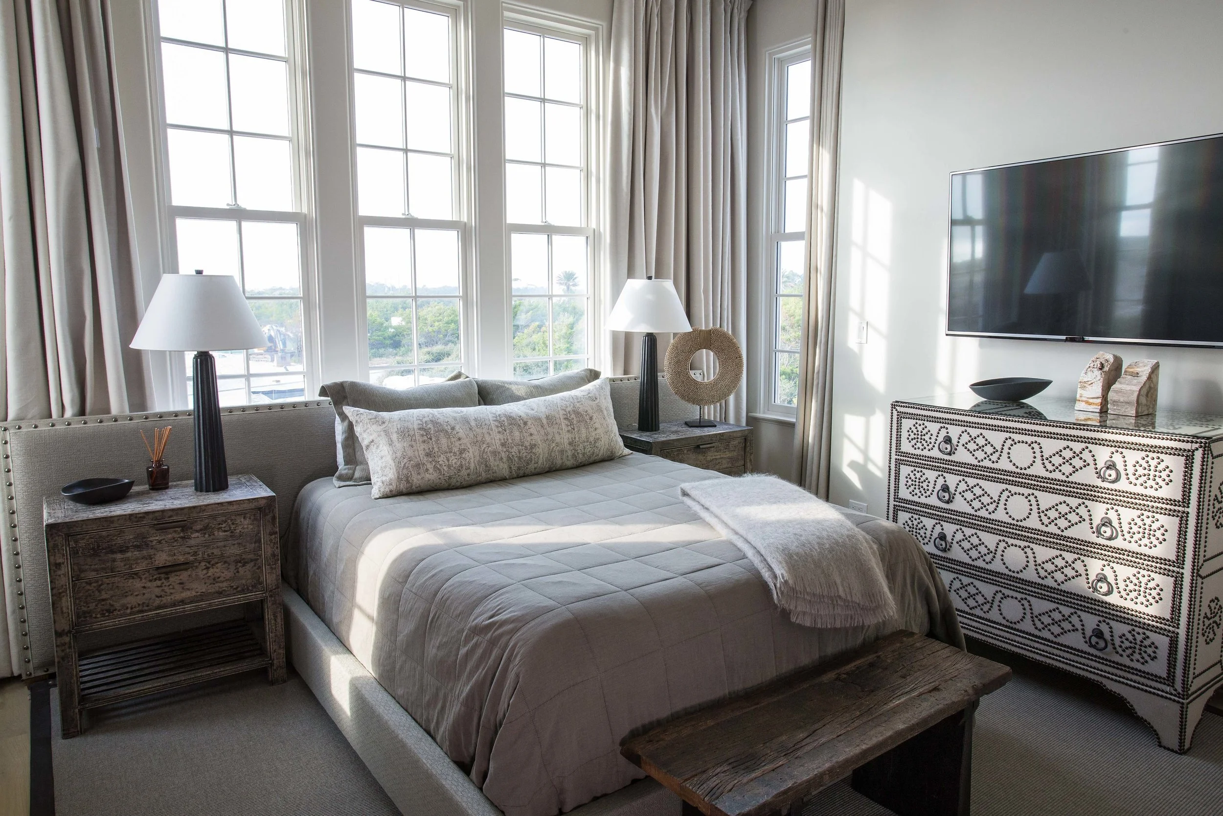 A cozy bedroom with large windows, a bed with beige bedding and pillows, two bedside tables with lamps, a decorative mirror and sculpture, and a dresser with a flat-screen TV mounted on the wall.