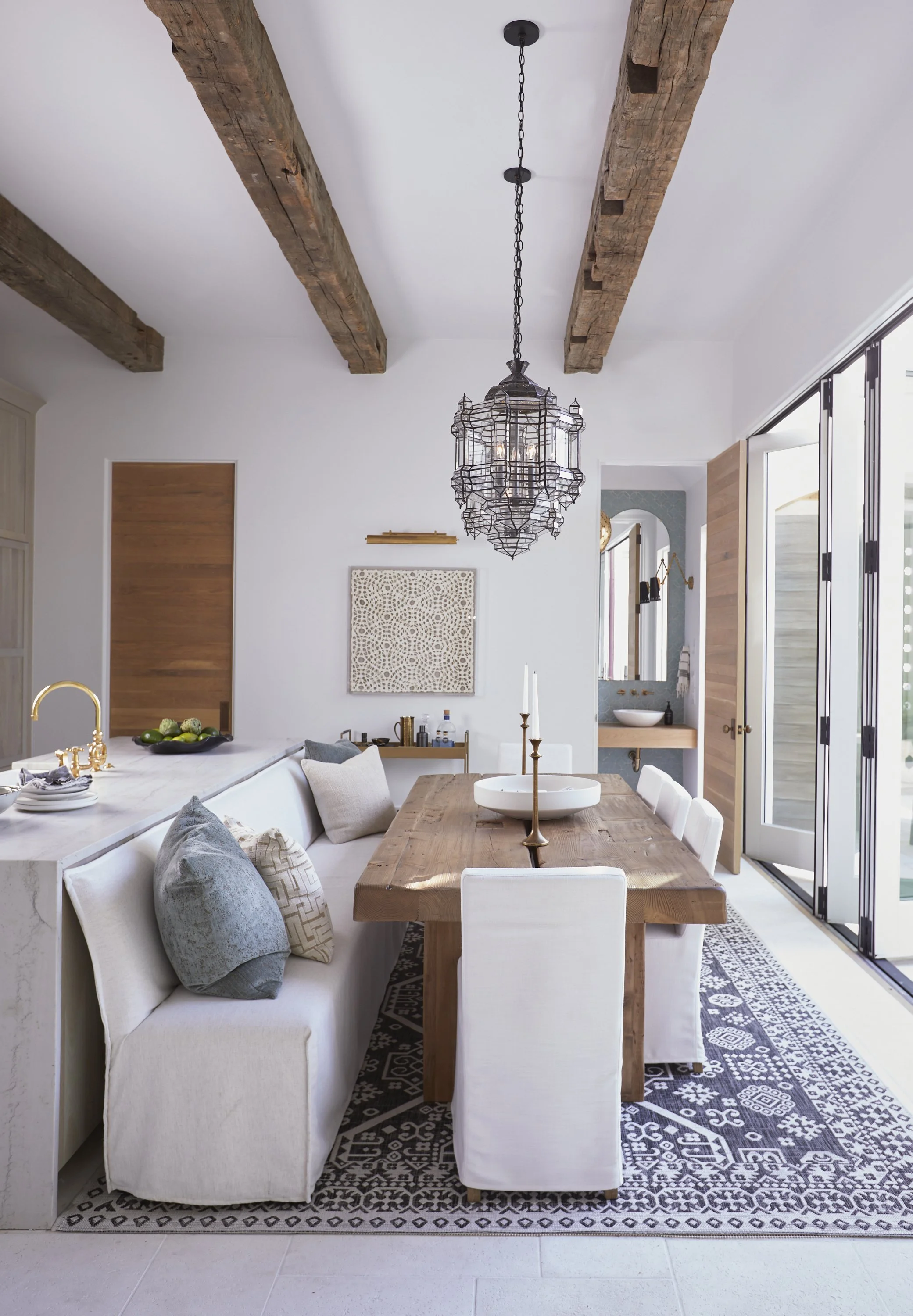 Modern dining room with a wooden table, white upholstered chairs, a white bench with pillows, a black patterned rug, a large black wrought iron chandelier, exposed wooden ceiling beams, and natural light coming through sliding glass doors.