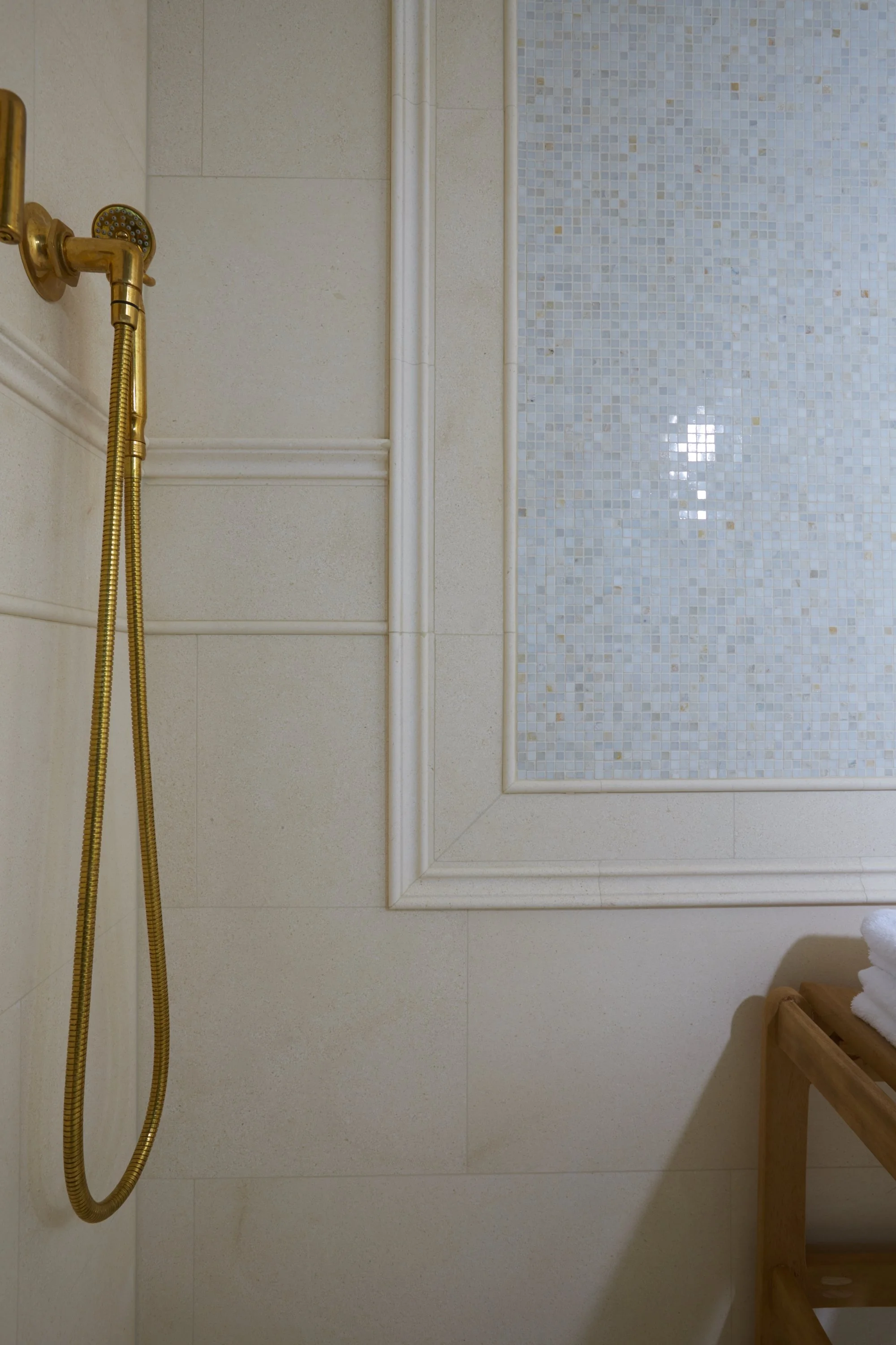Close-up of a shower area with a gold-colored shower head and hose attached to a wall. The wall has beige tiles and a decorative frame surrounding a mosaic tile panel. A wooden shelf with folded white towels is partially visible in the bottom right c