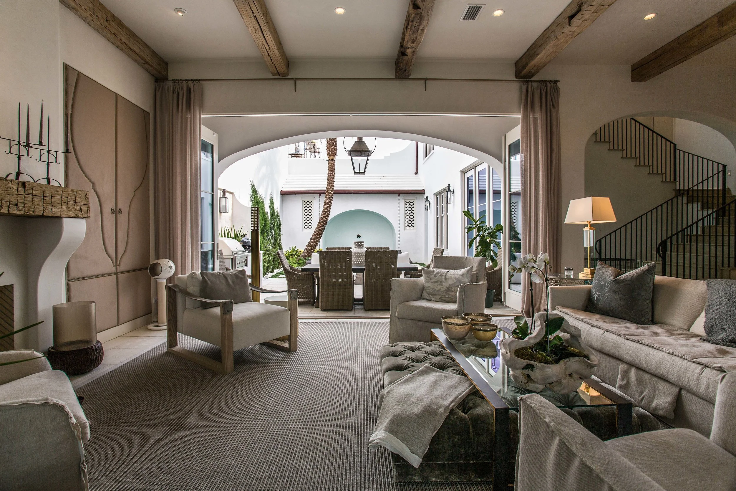 Living room with beige sofas, armchairs, a coffee table, and an open door revealing an outdoor patio with dining furniture, centered around a palm tree.