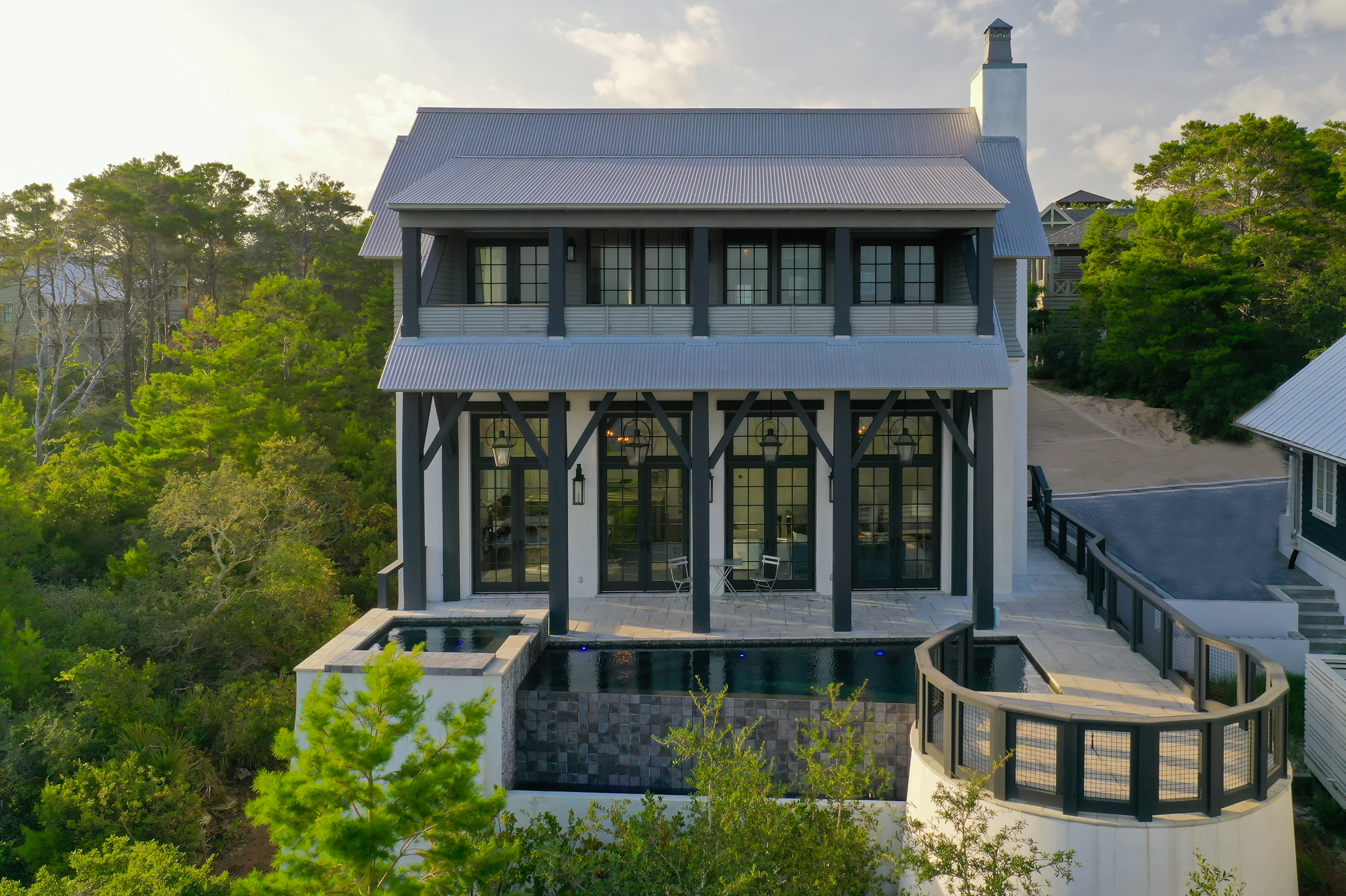 Coastal house with black trim, multiple balconies, and a rooftop patio overlooking trees and ocean in the background.