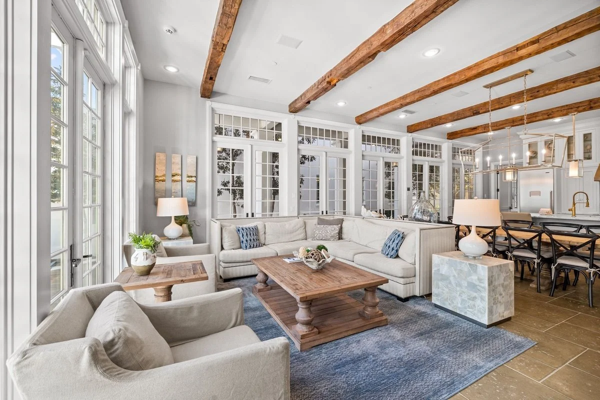 Bright living room with large windows, white walls, white furniture, wooden ceiling beams, and a kitchen area in the background.