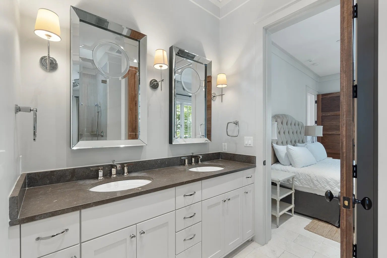 Bathroom vanity with two sinks, mirrors, wall-mounted lights, and a view into a bedroom with a bed and nightstand.