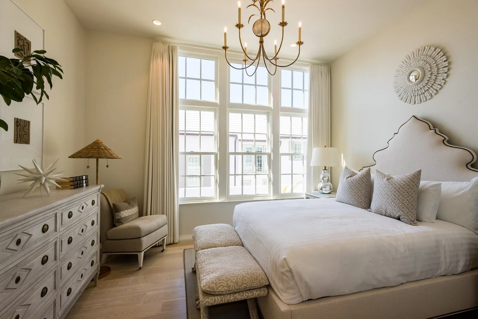 A cozy bedroom with large windows, beige furniture, a chandelier, and elegant decor.