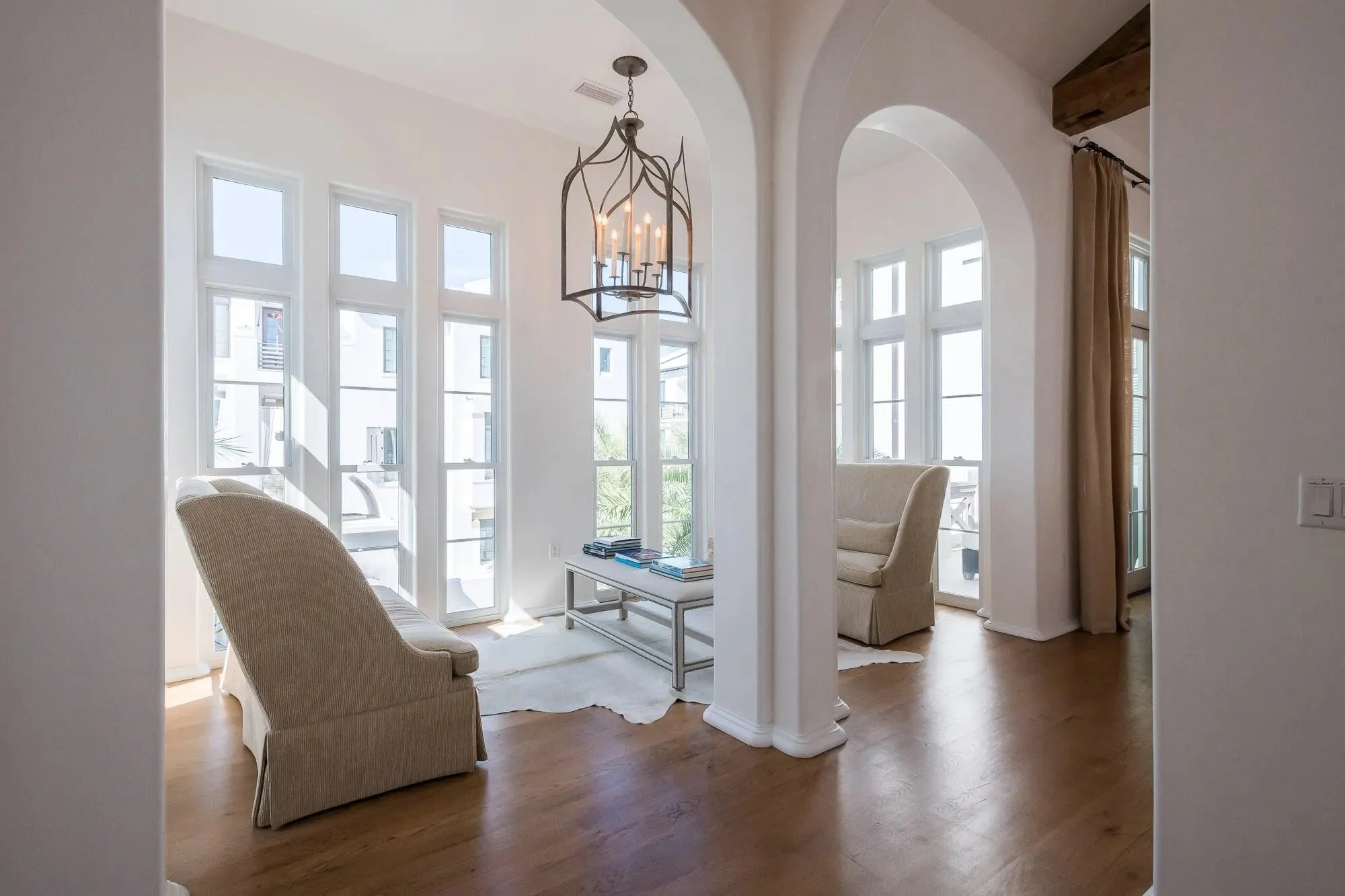 Bright living room with large windows, white walls, beige armchairs, a glass coffee table with books, hardwood floors, and a decorative chandelier hanging from the ceiling.
