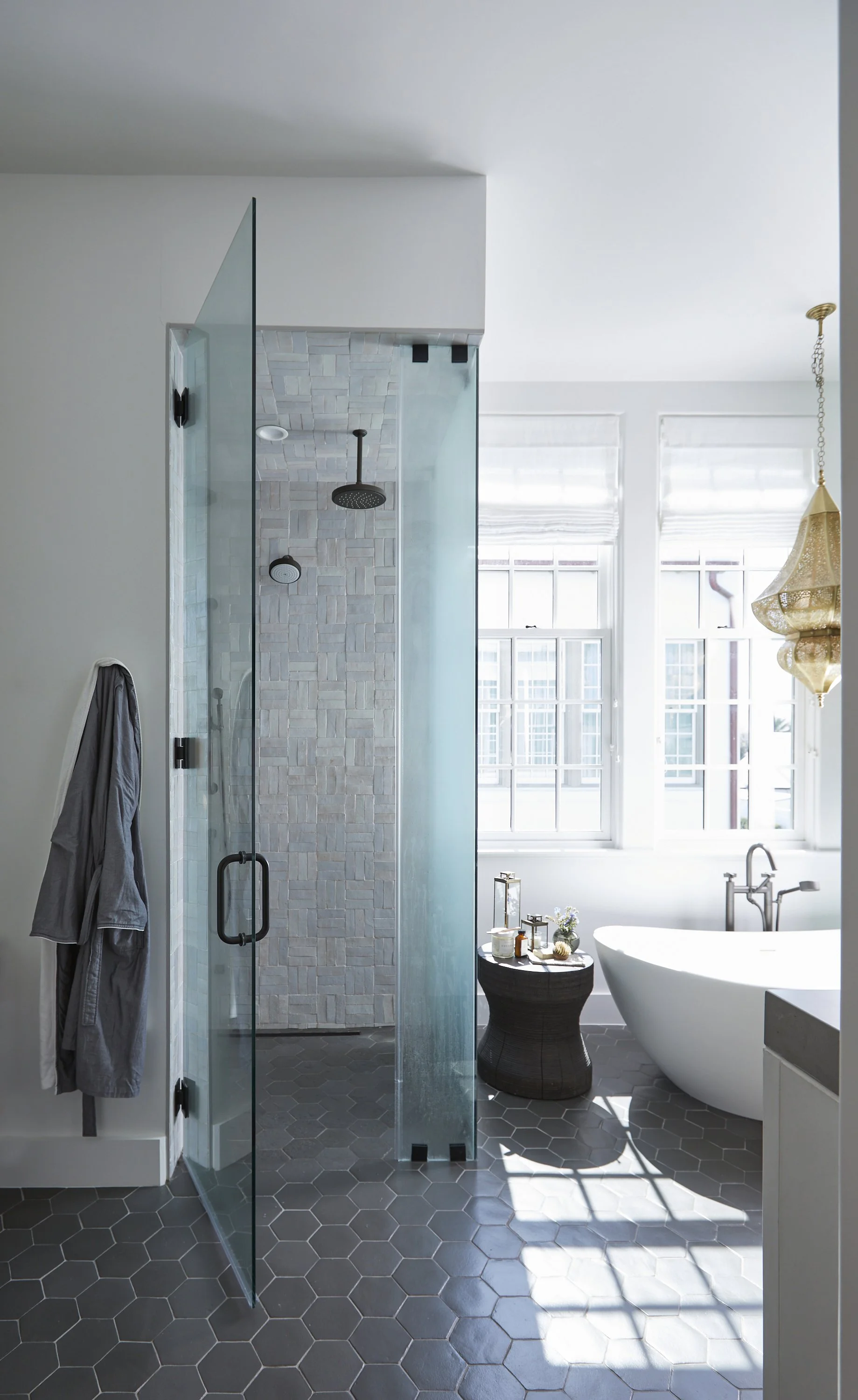 Modern bathroom with a glass shower enclosure, a freestanding bathtub by windows, and a pendant chandelier.