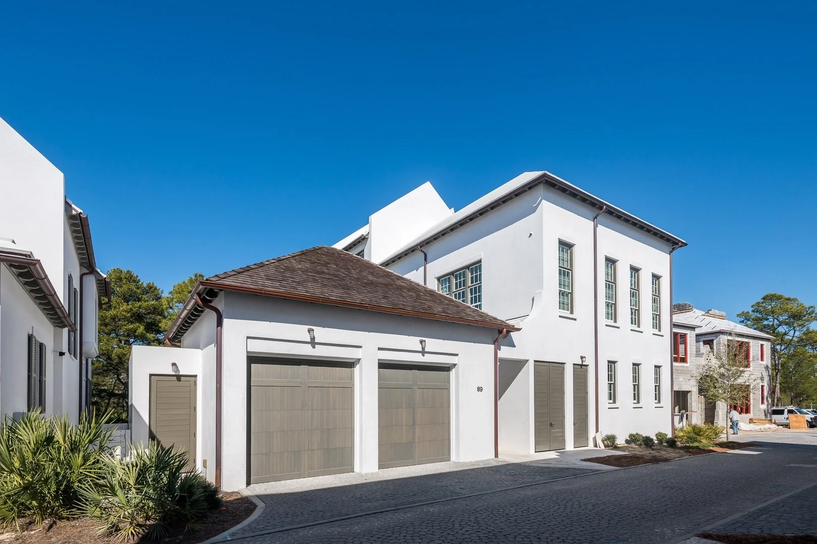 Modern white multi-story residential buildings with garage doors, clear blue sky, some trees, and landscaped surroundings.