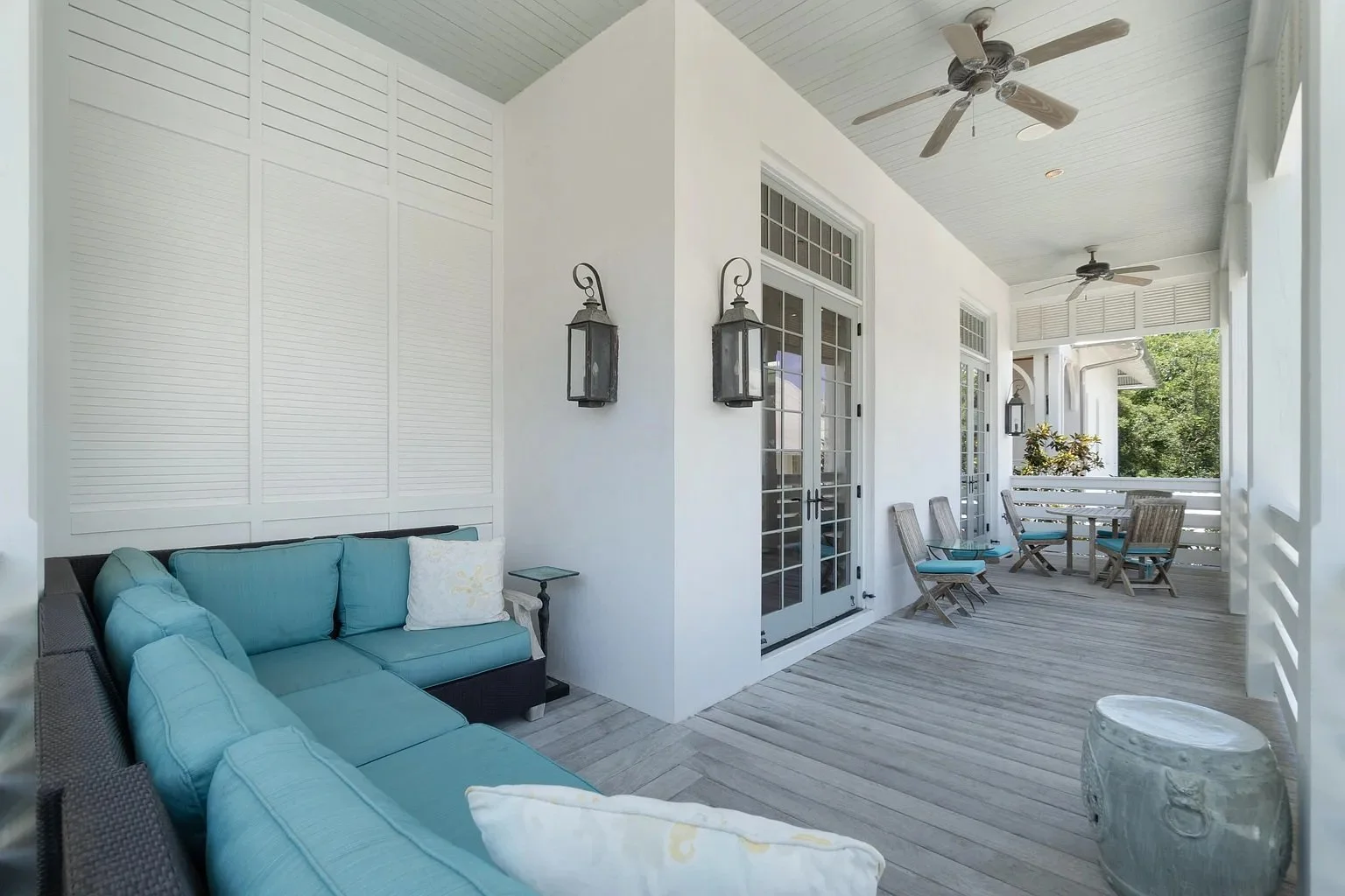 Covered porch with light blue outdoor sofa, wicker chairs with cushions, and a round table. White walls with French doors, lantern-style wall sconces, ceiling fans, and a wooden floor with a view of greenery outside.