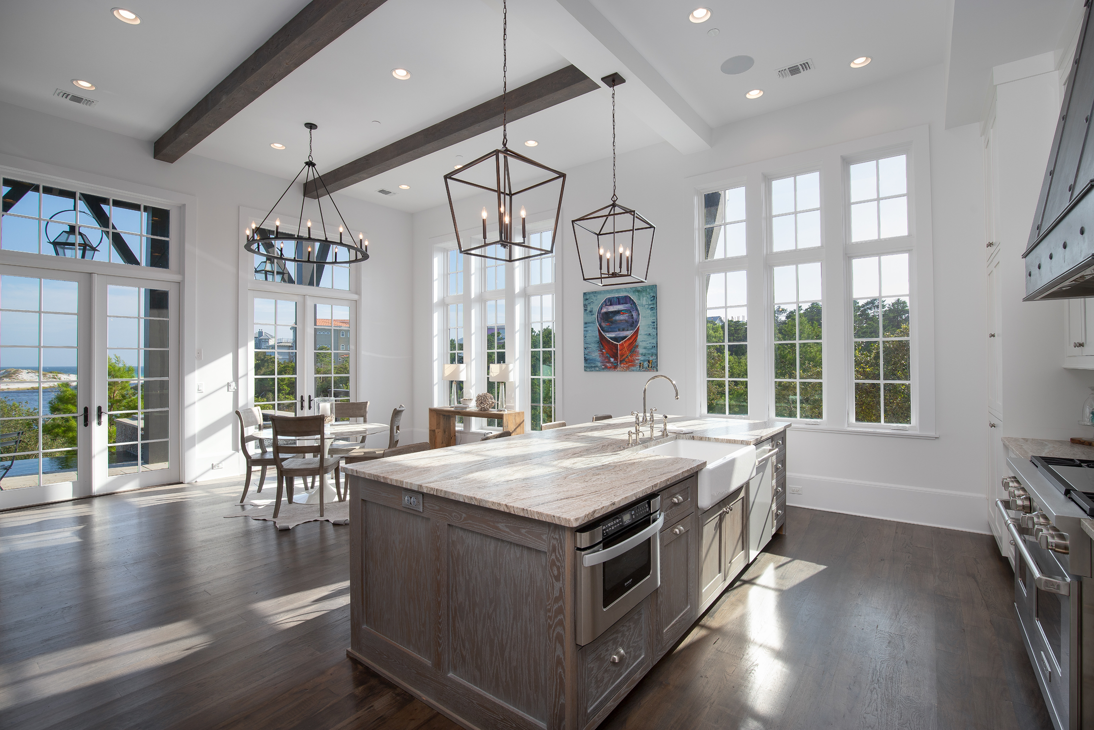 Bright open kitchen with large windows, a wooden island with a sink, pendant lights, a dining area with a round table and chairs, and outdoor scenery visible through the windows and glass doors.